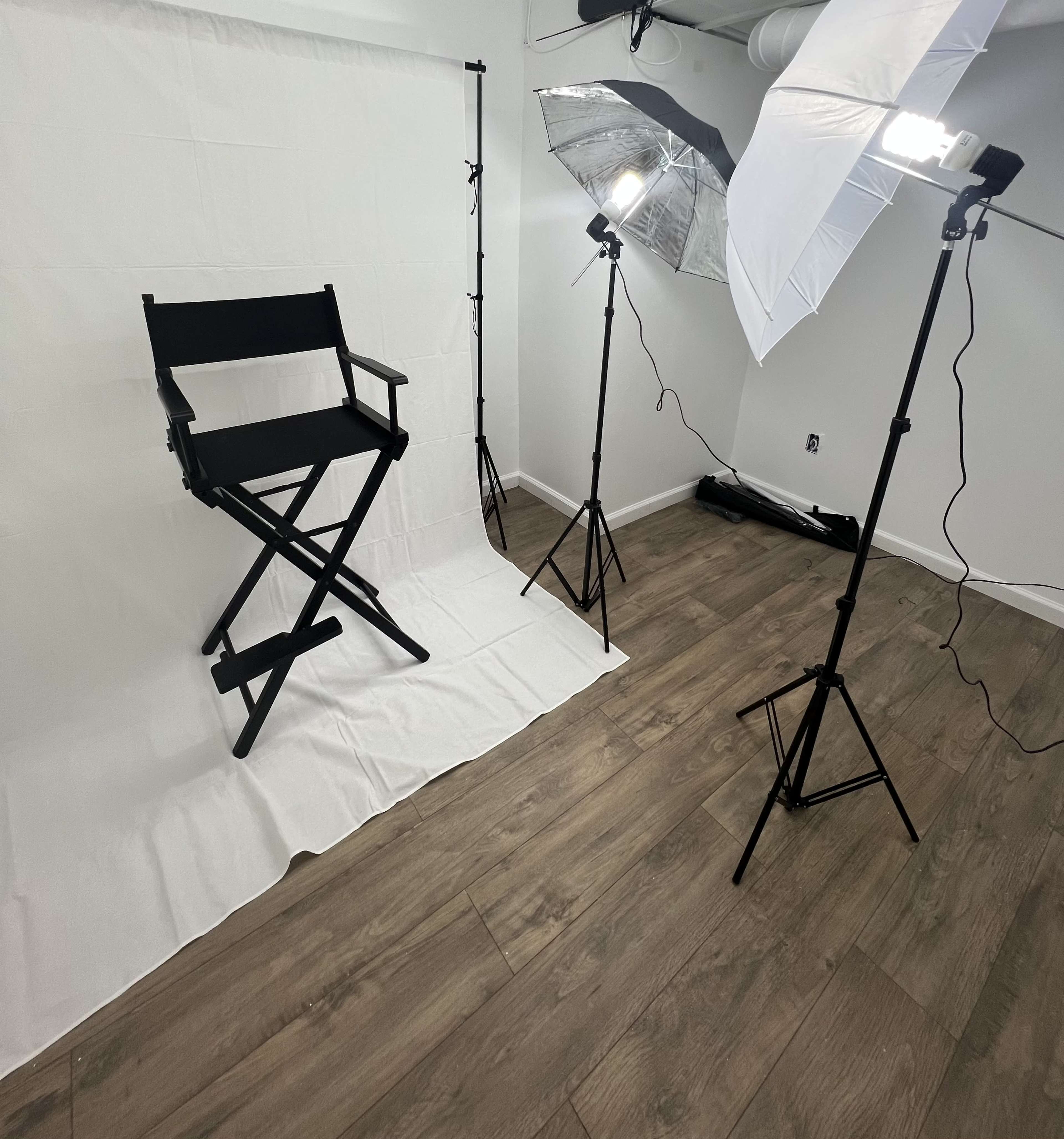 A photography setup with a black director's chair on a white backdrop, flanked by two light stands with softboxes, positioned on a wooden floor.