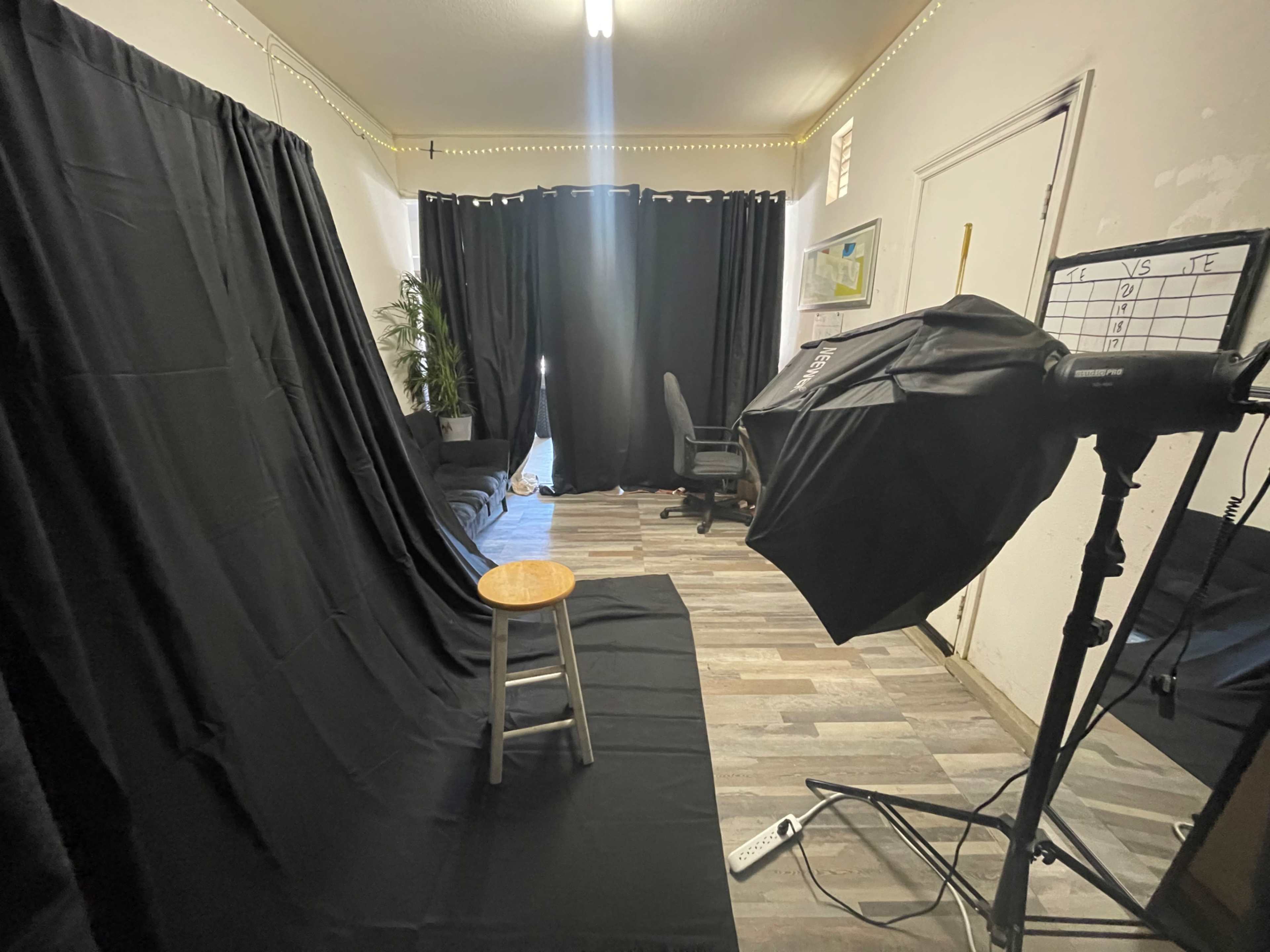 A photography setup with a black backdrop, a wooden stool in the center, and a lighting equipment on the side, all located in a room with dark curtains and minimal furniture.