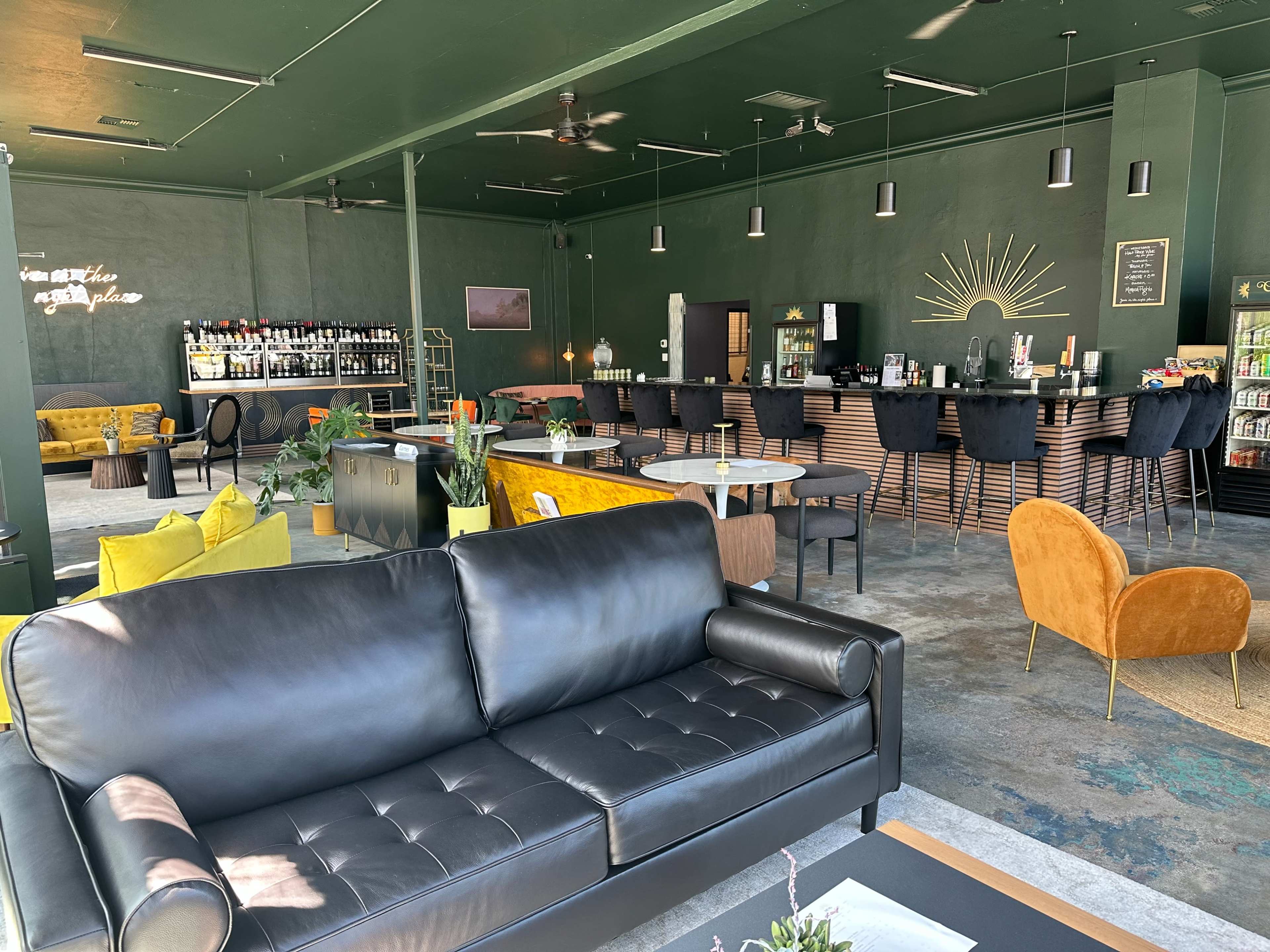 The image shows a stylish lounge area featuring modern seating, a bar counter with various bottles, and vibrant upholstery in a green-toned space.