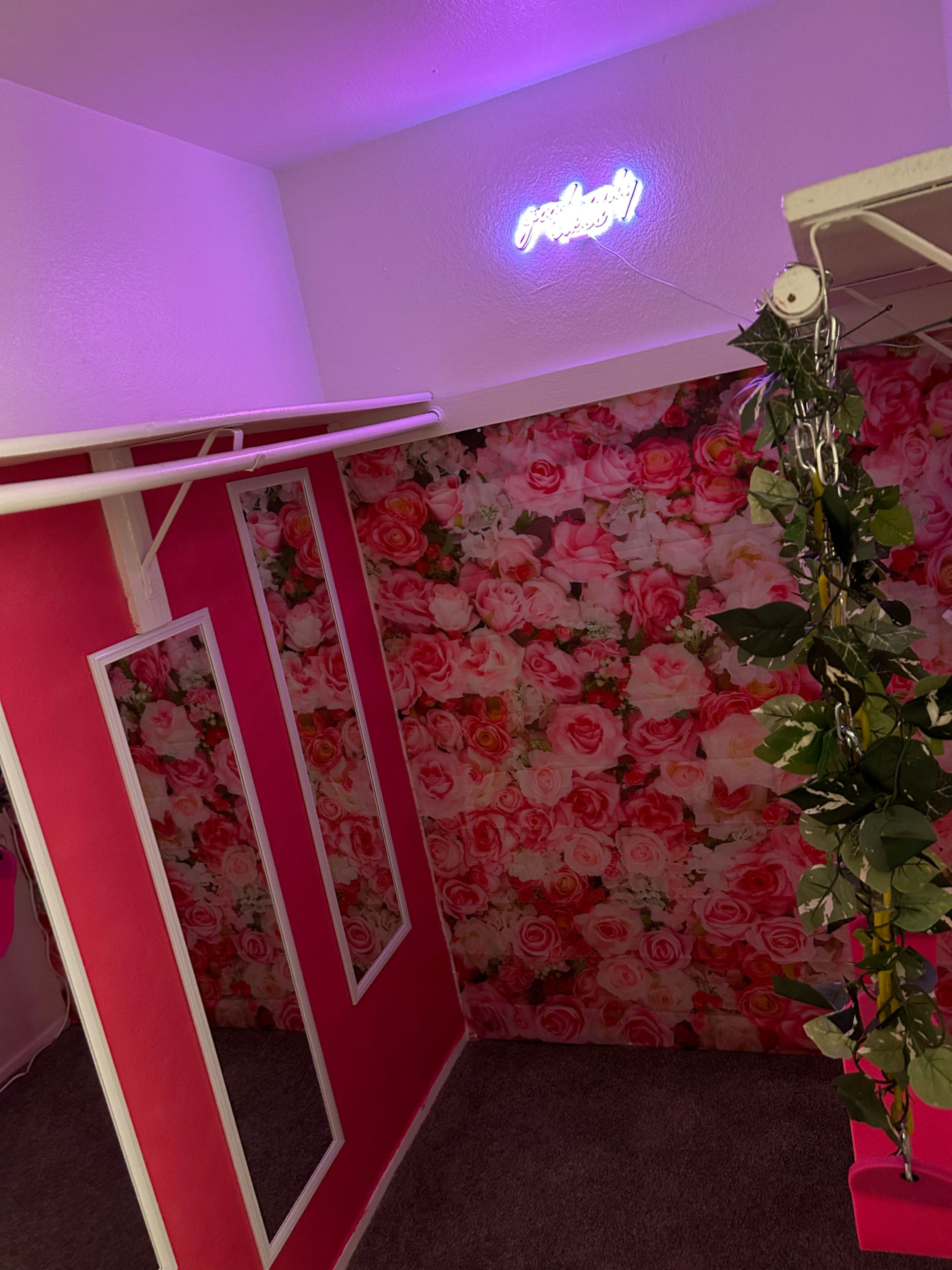 The image shows a corner of a room with pink walls, a floral-patterned background, and a neon sign that says "Perfectly."