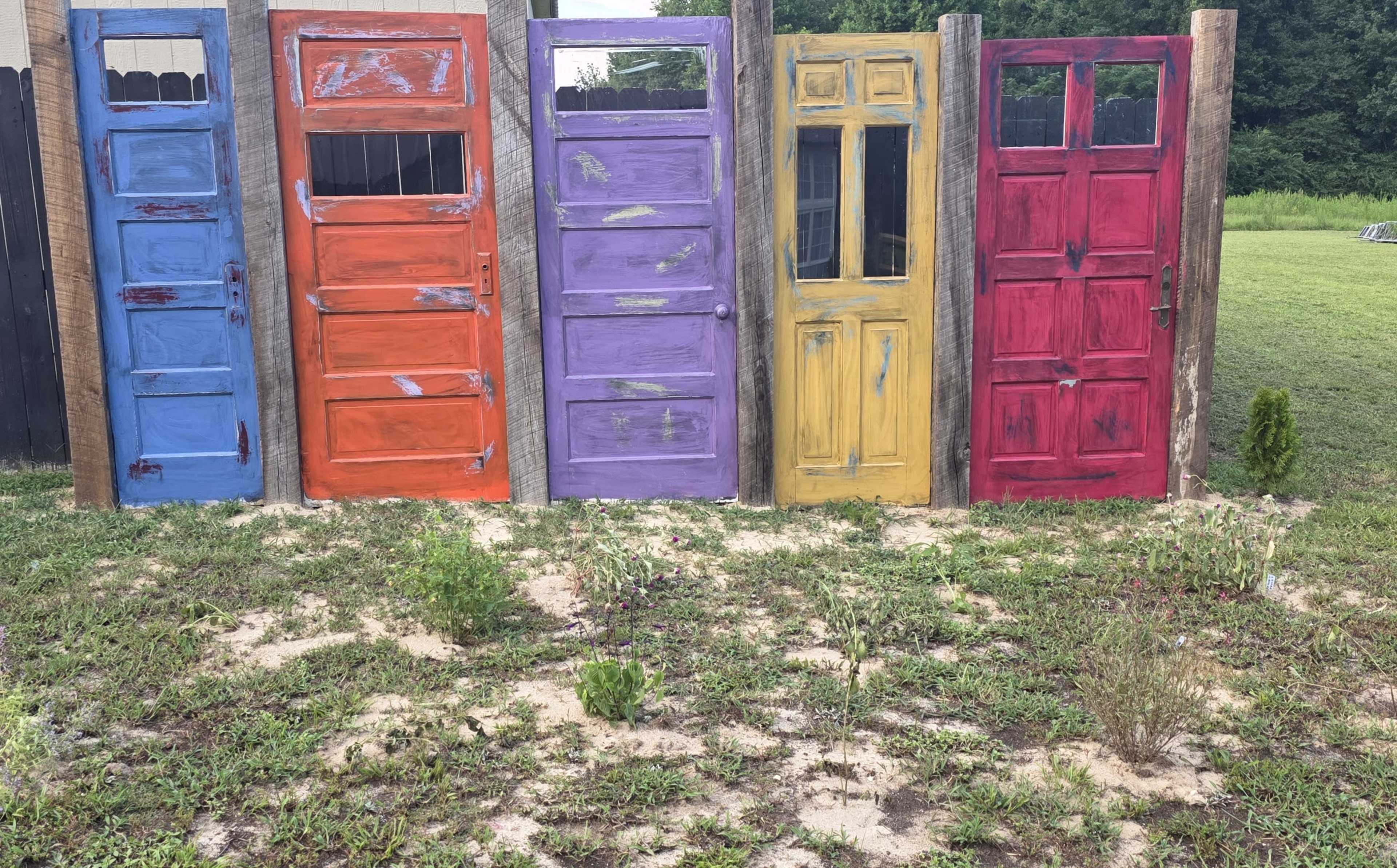 The image shows a row of five colorful doors painted in orange, purple, yellow, red, and blue, set against a grassy landscape.