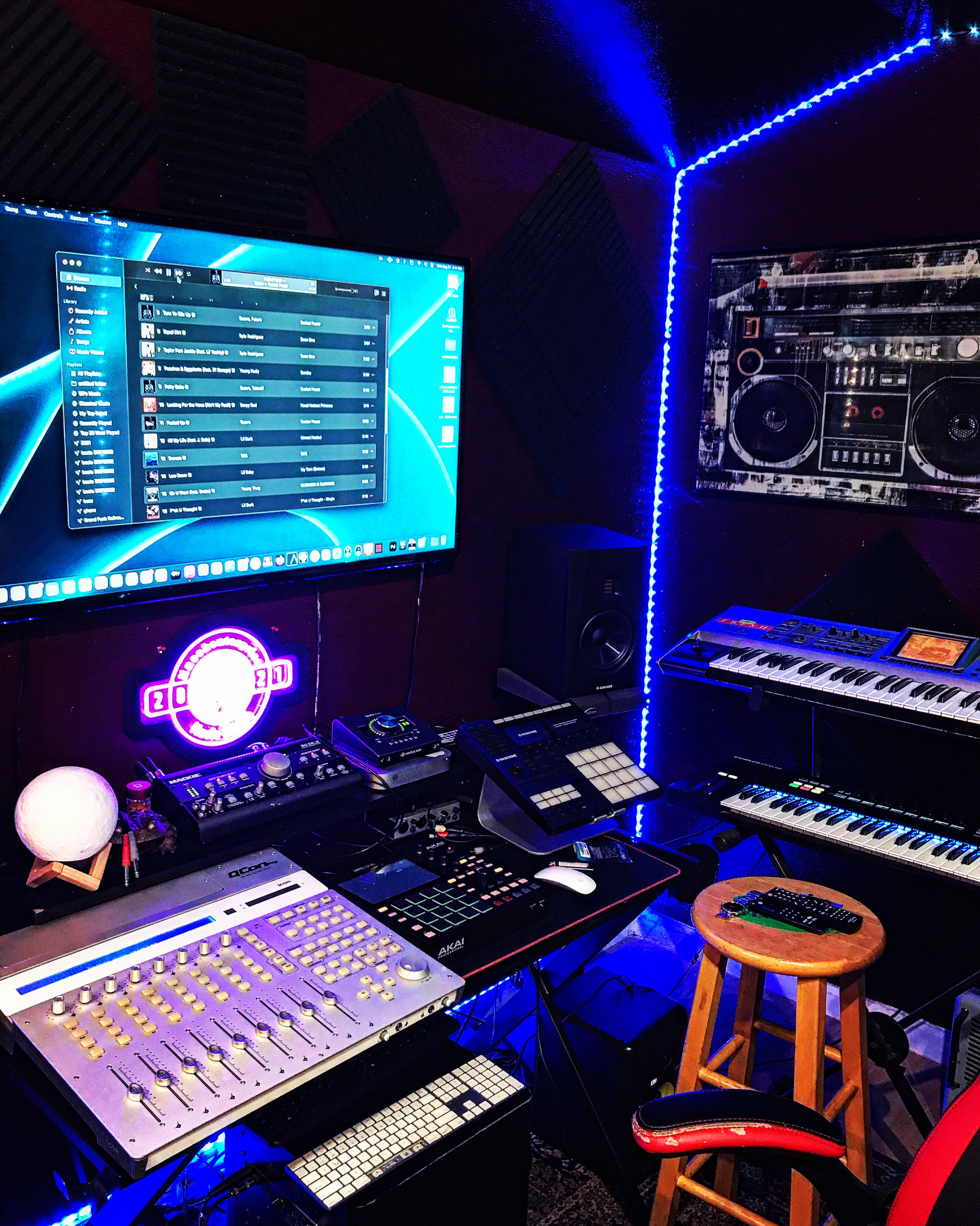 A music production studio filled with various electronic equipment, including a computer monitor displaying software, a keyboard, and a mixing console, with LED lights illuminating the space.
