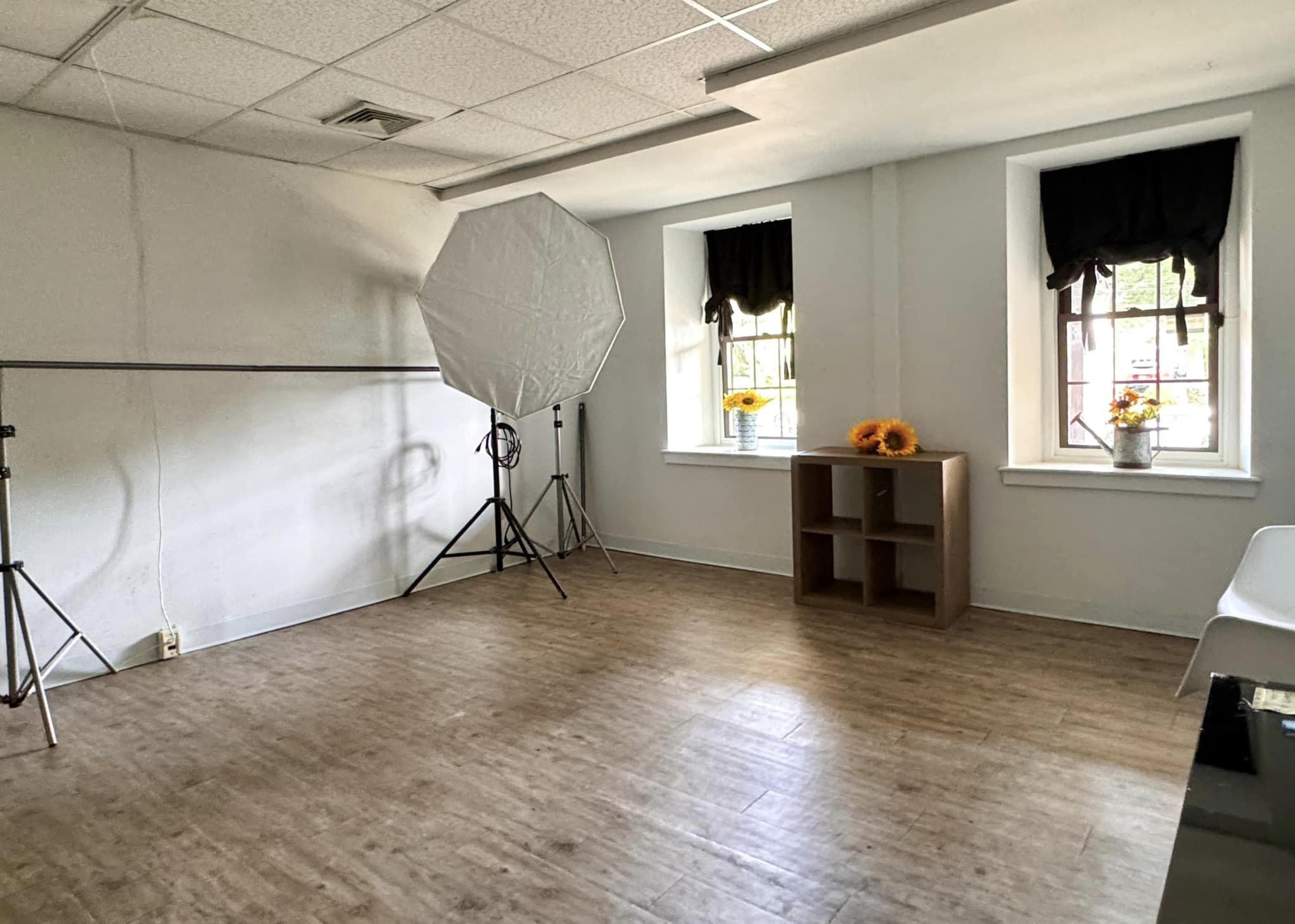 The image shows a vacant room with two windows, a light-colored floor, a photo backdrop setup, a small shelving unit, and a chair.