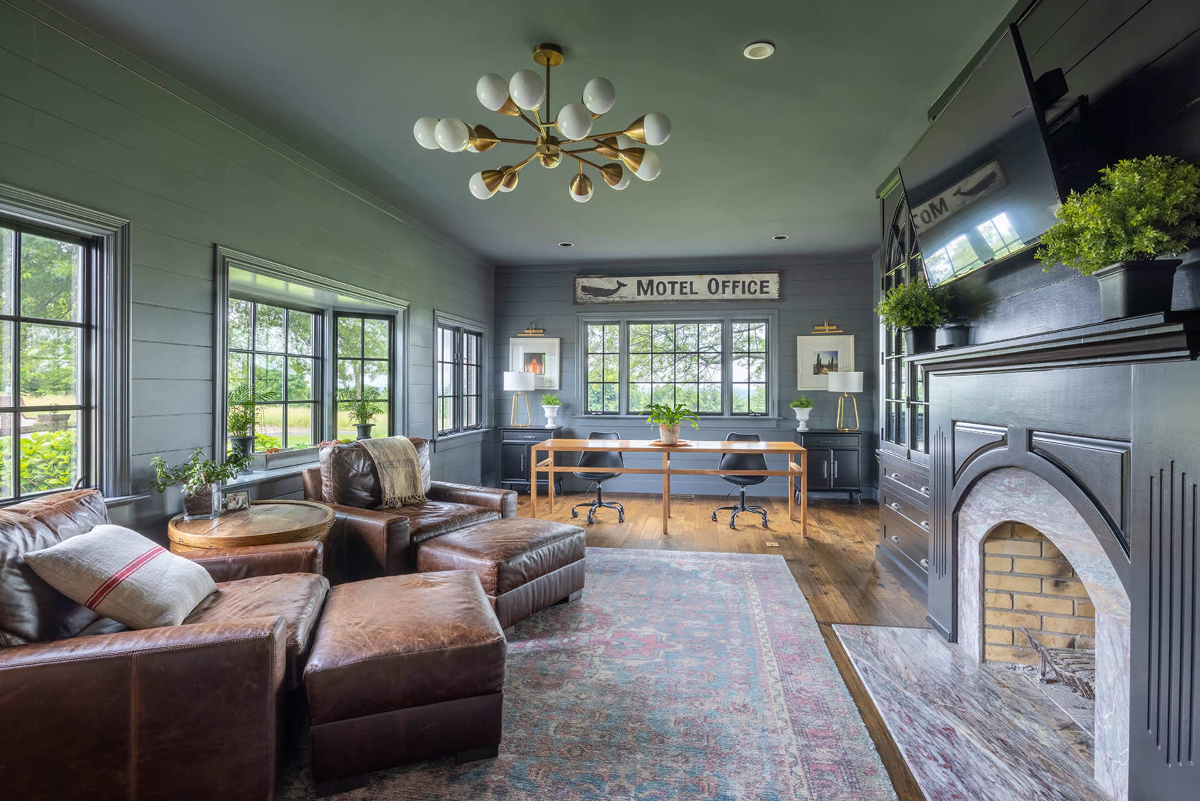 The image shows a motel office interior featuring a seating area with brown leather chairs, a wooden desk, and large windows allowing natural light to enter.