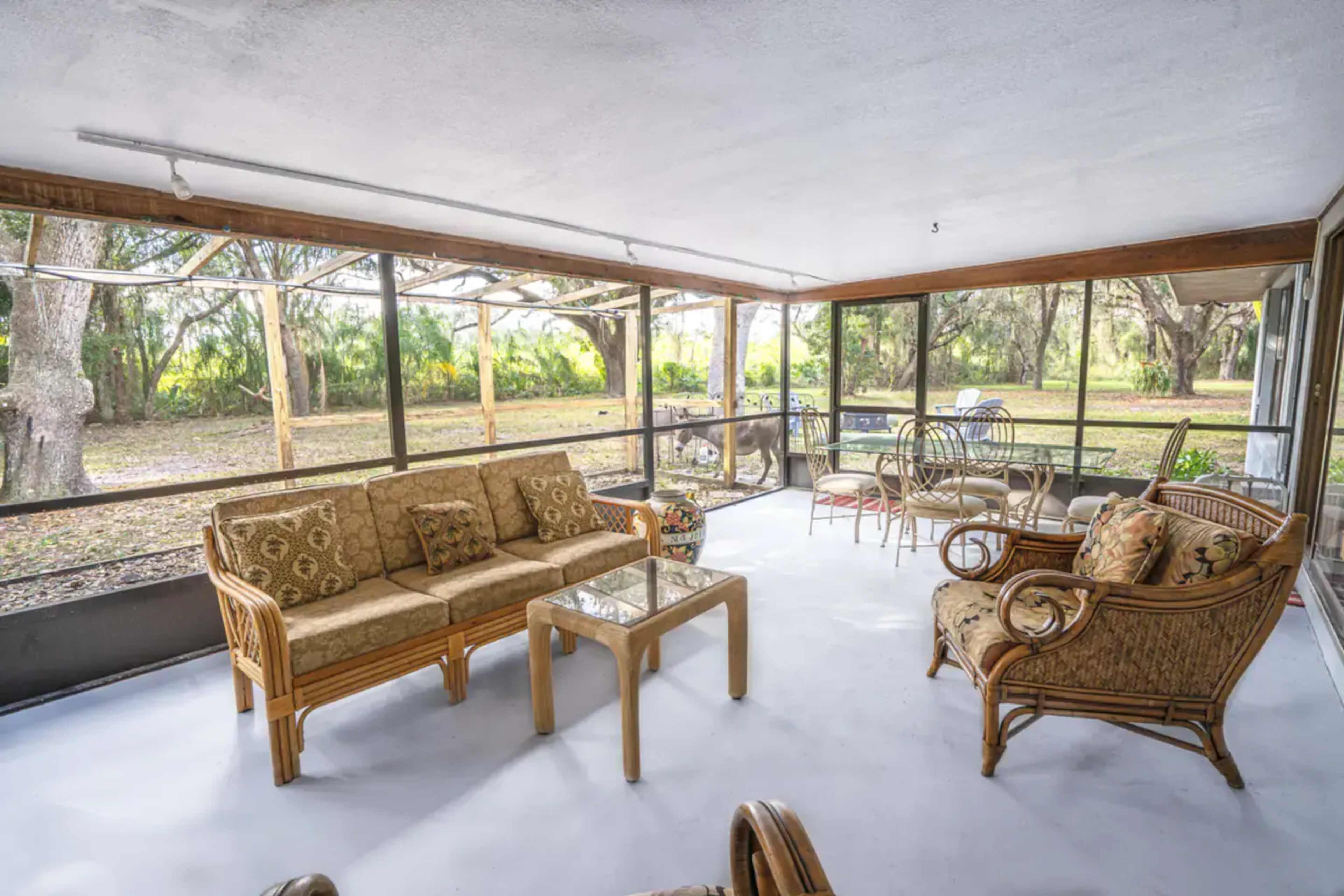 The image shows a sunroom with wicker furniture, including a sofa, armchairs, and a glass coffee table, overlooking a green outdoor area through large screened windows.
