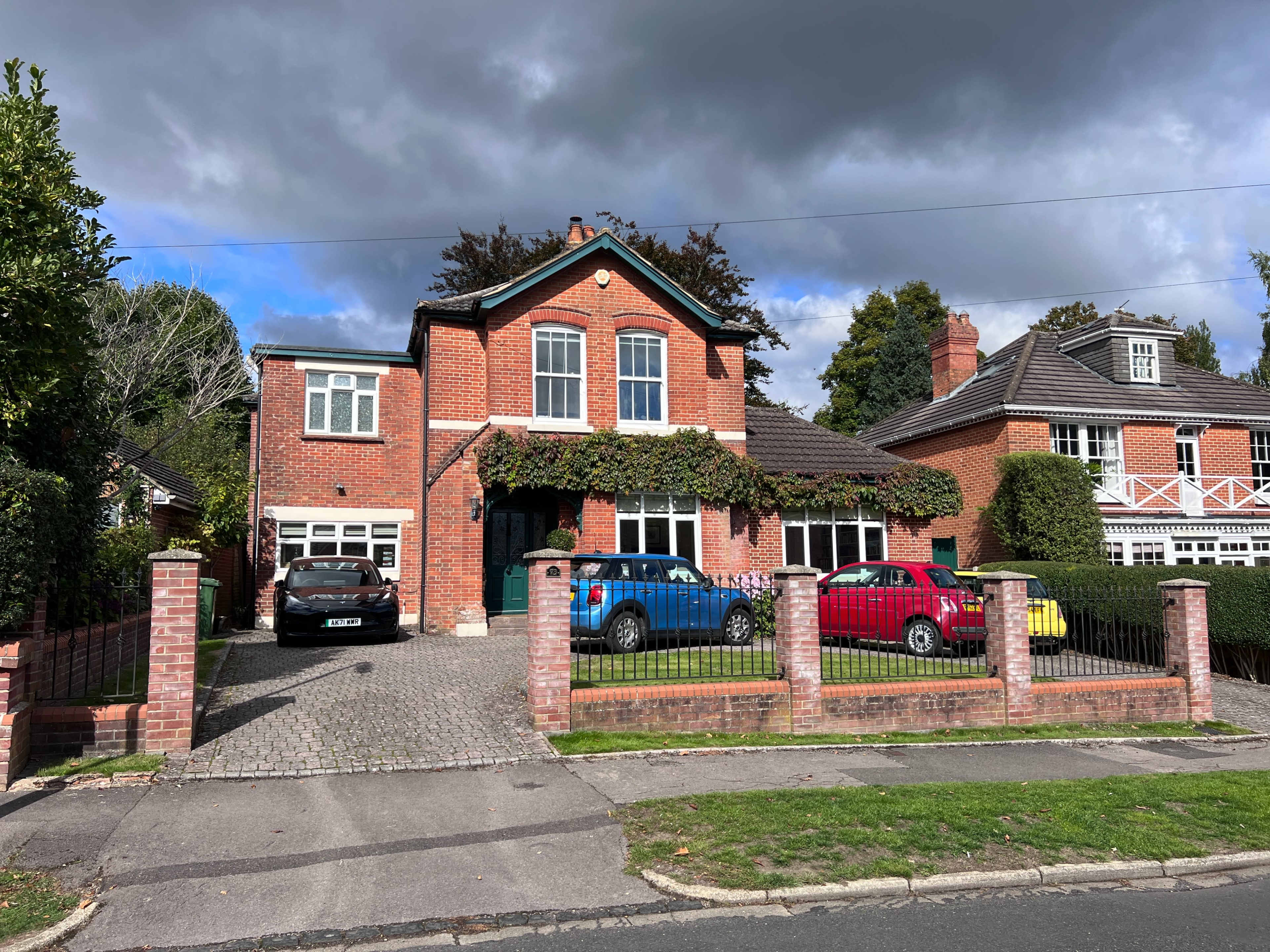 The photograph shows a two-story brick house with a driveway containing four parked cars in front of a well-manicured yard and a cloudy sky above.