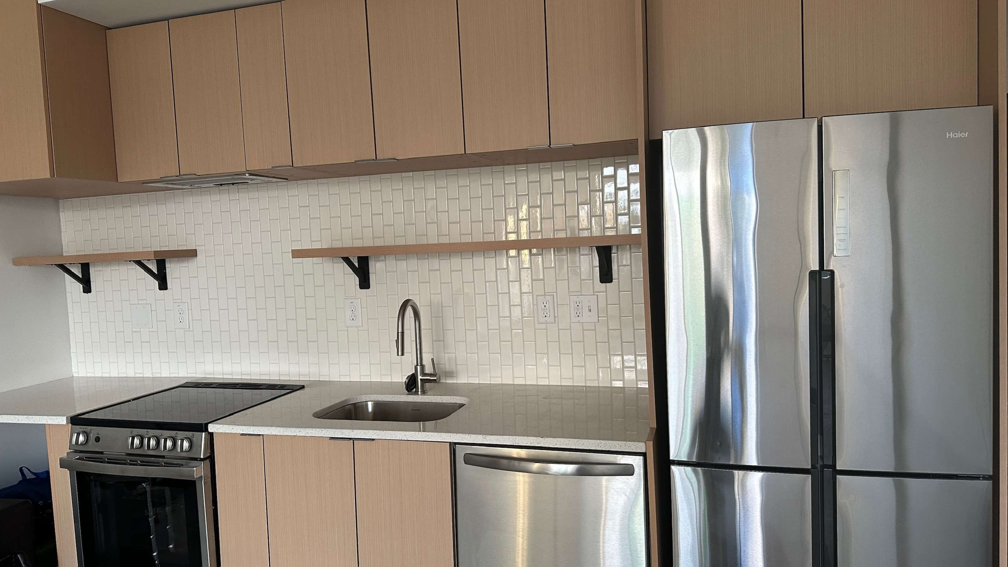 A modern kitchen with wooden cabinets, a stainless steel refrigerator, an oven, and a sink, set against a white tile backsplash.