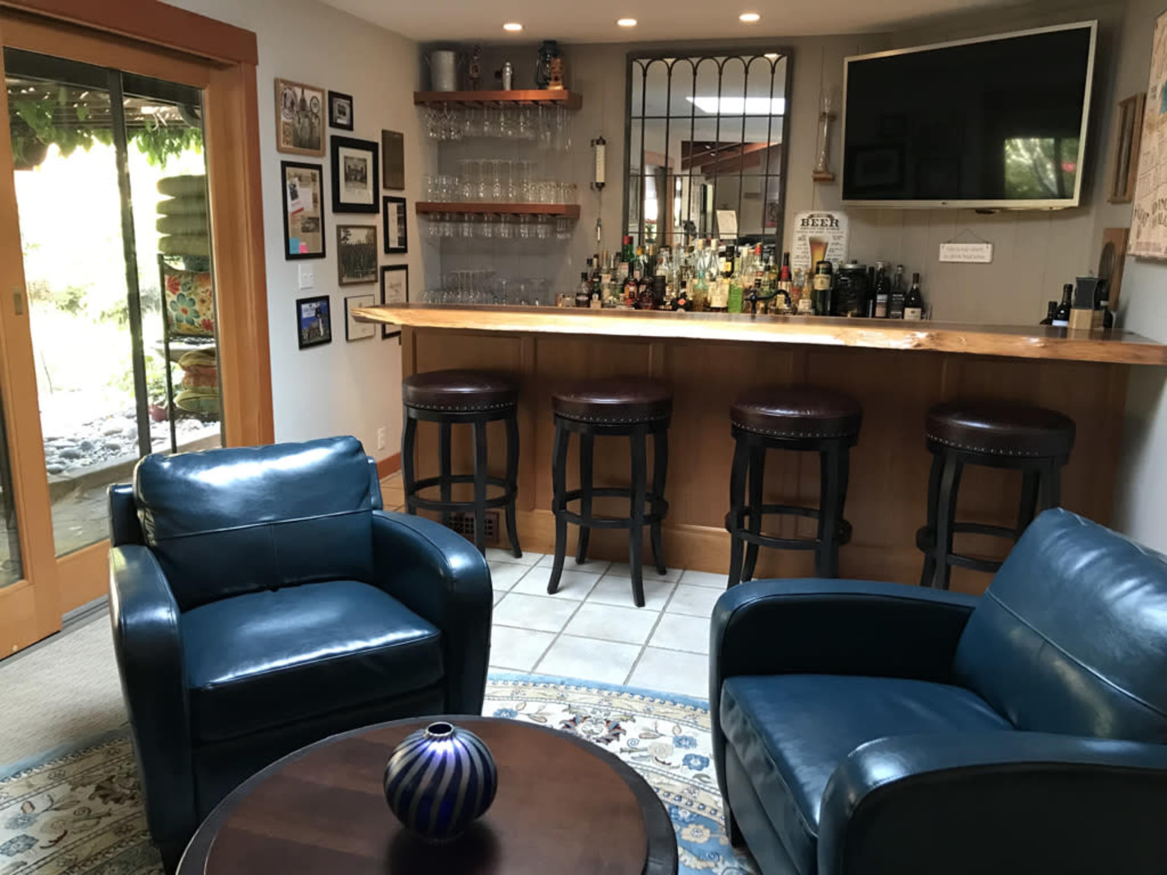 The scene shows a cozy home bar area with two blue leather chairs facing a wooden bar with several bottles and glasses behind it, and a television mounted on the wall.