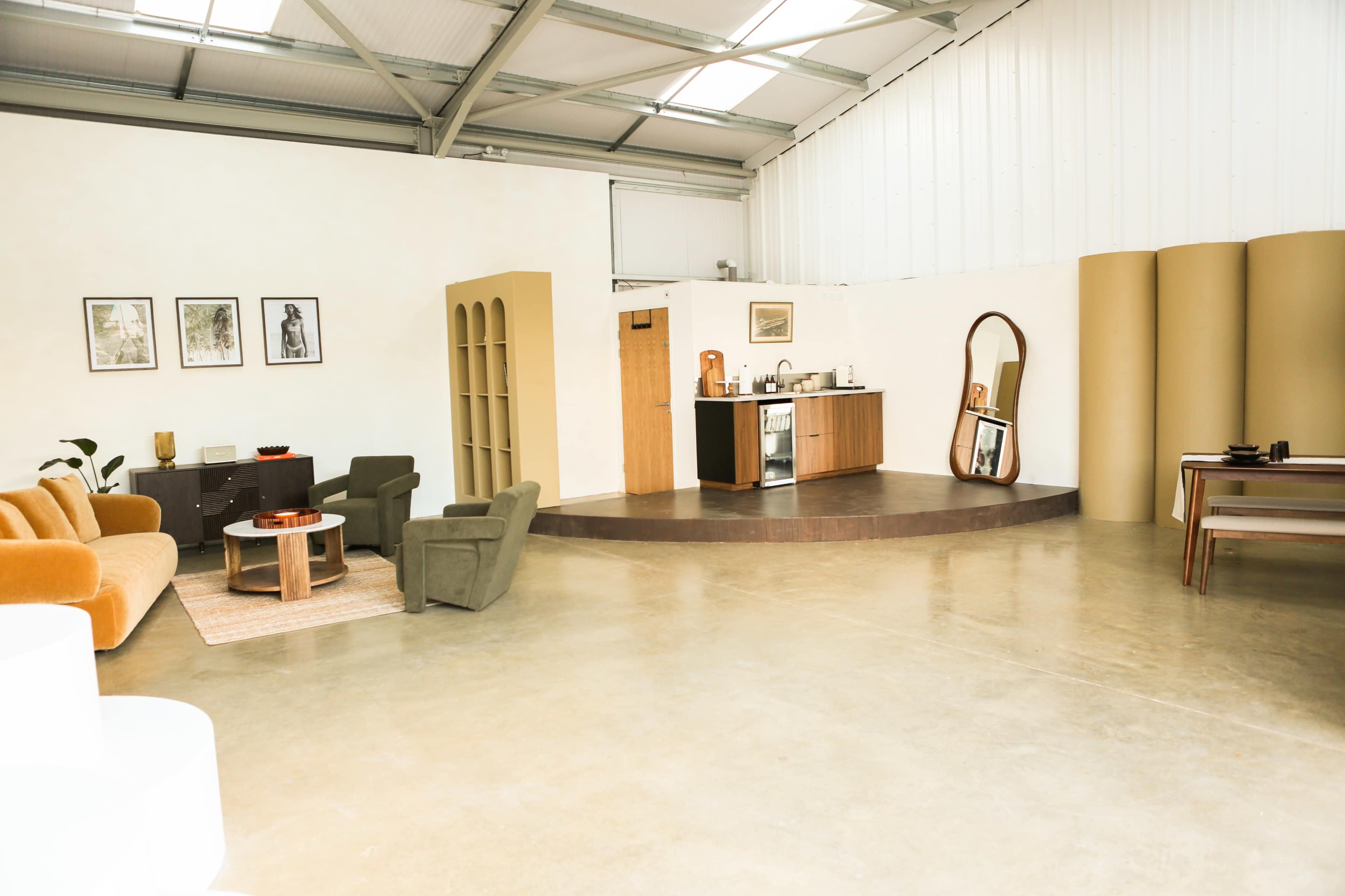 The image shows a spacious, modern interior with a kitchenette, seating area, and large windows.