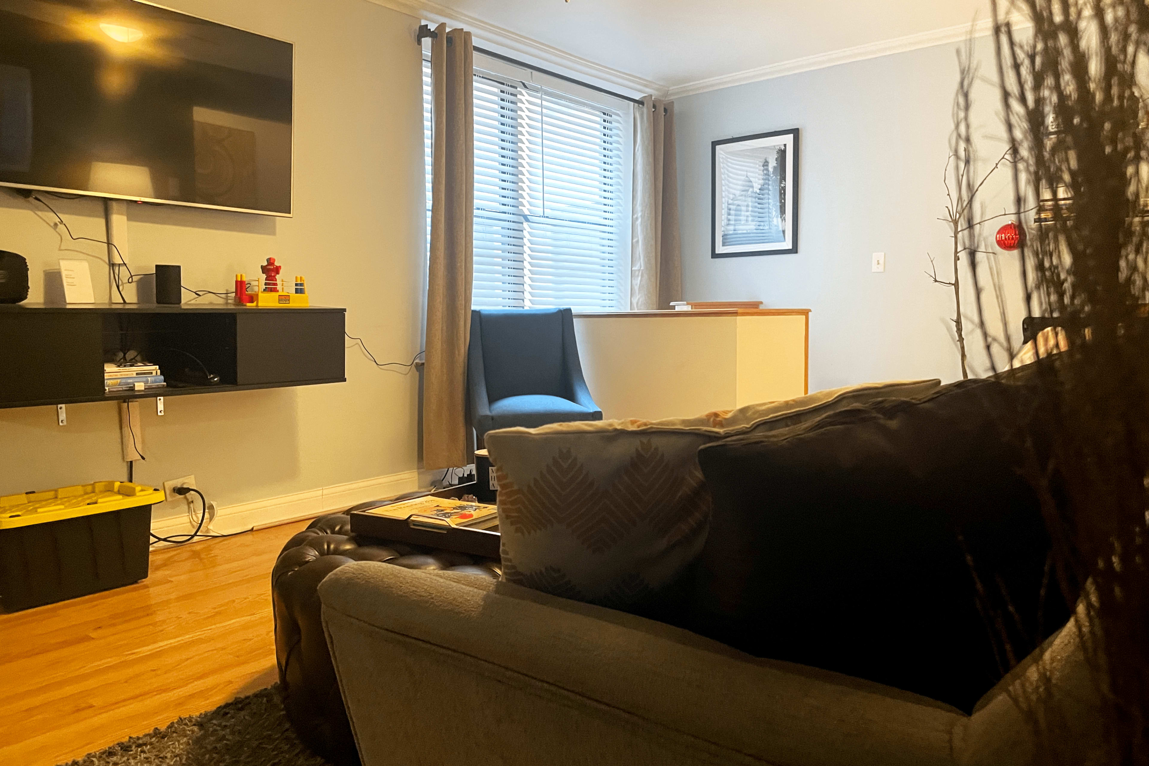 A cozy living room features a gray couch, a coffee table, a television mounted on the wall, and a small desk with a chair near a window.
