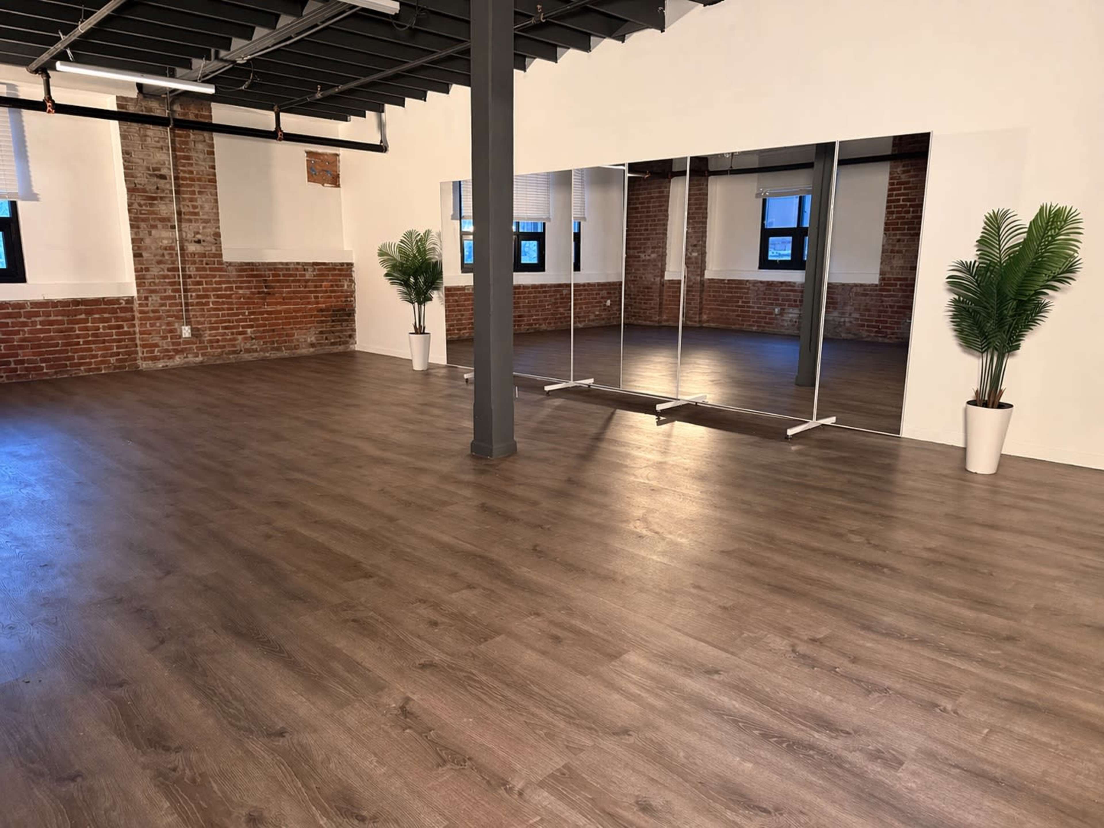The image shows a spacious room with wooden flooring, exposed brick walls, large mirrors, and potted plants in the corners.