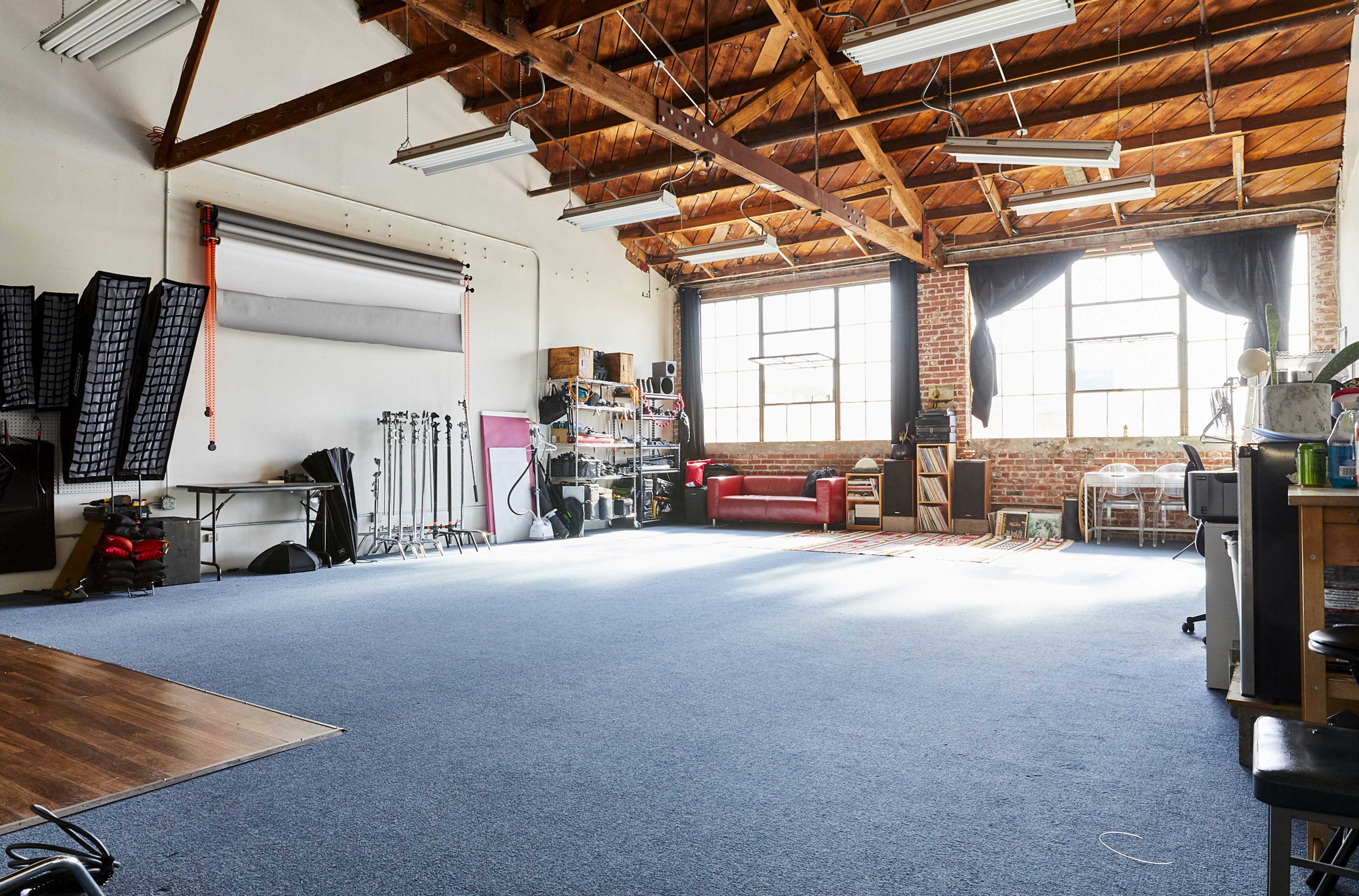 The image shows a spacious studio with wooden beams, large windows, a red sofa, and various equipment along the walls.