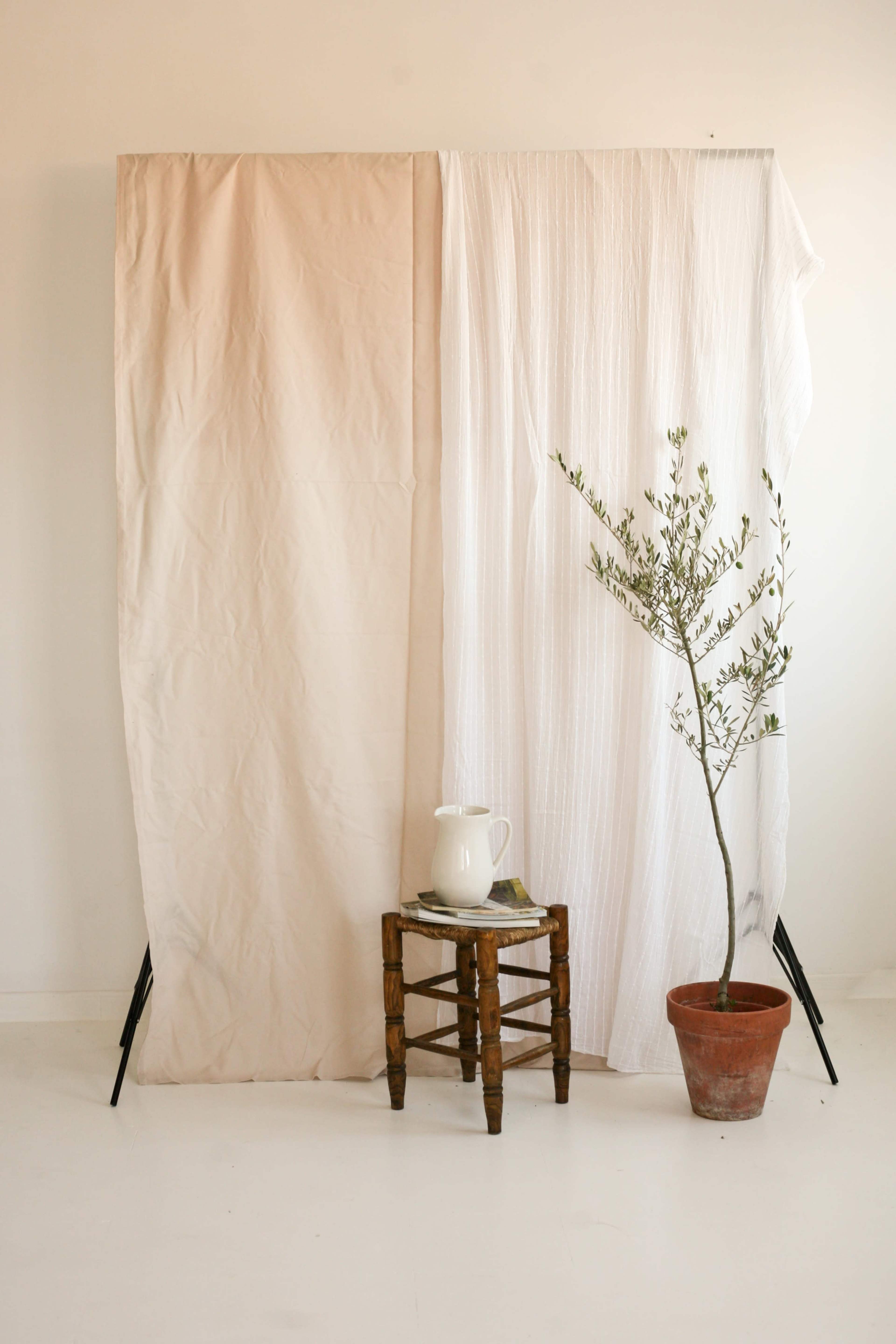 A minimalistic interior scene with a wooden stool, a potted plant, and a pitcher on a small table, separated by two draped fabric panels.