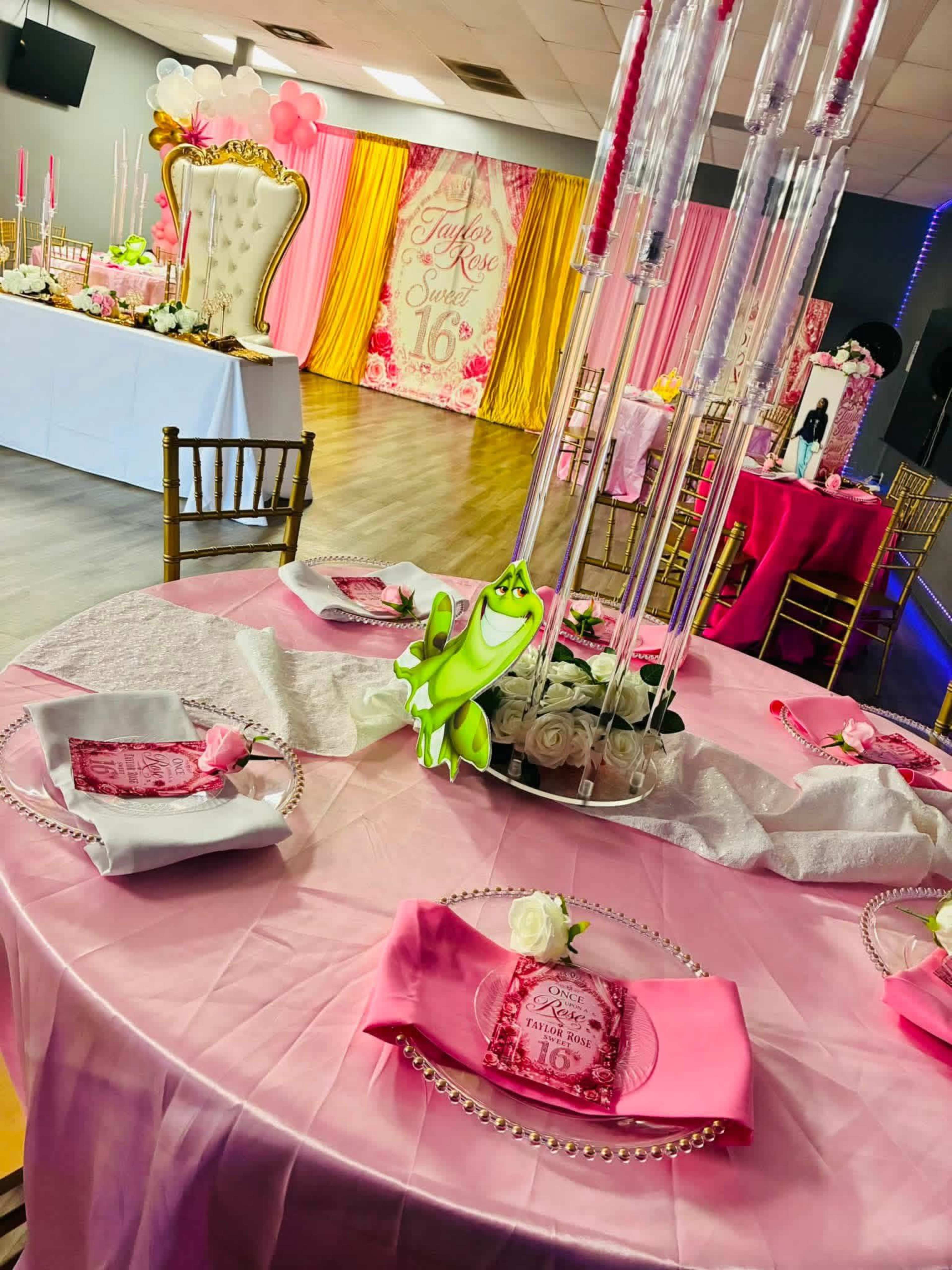 The image shows a decorated event space featuring tables set with pink and white tablecloths, elegant centerpieces, and a backdrop adorned with floral designs for a celebration.