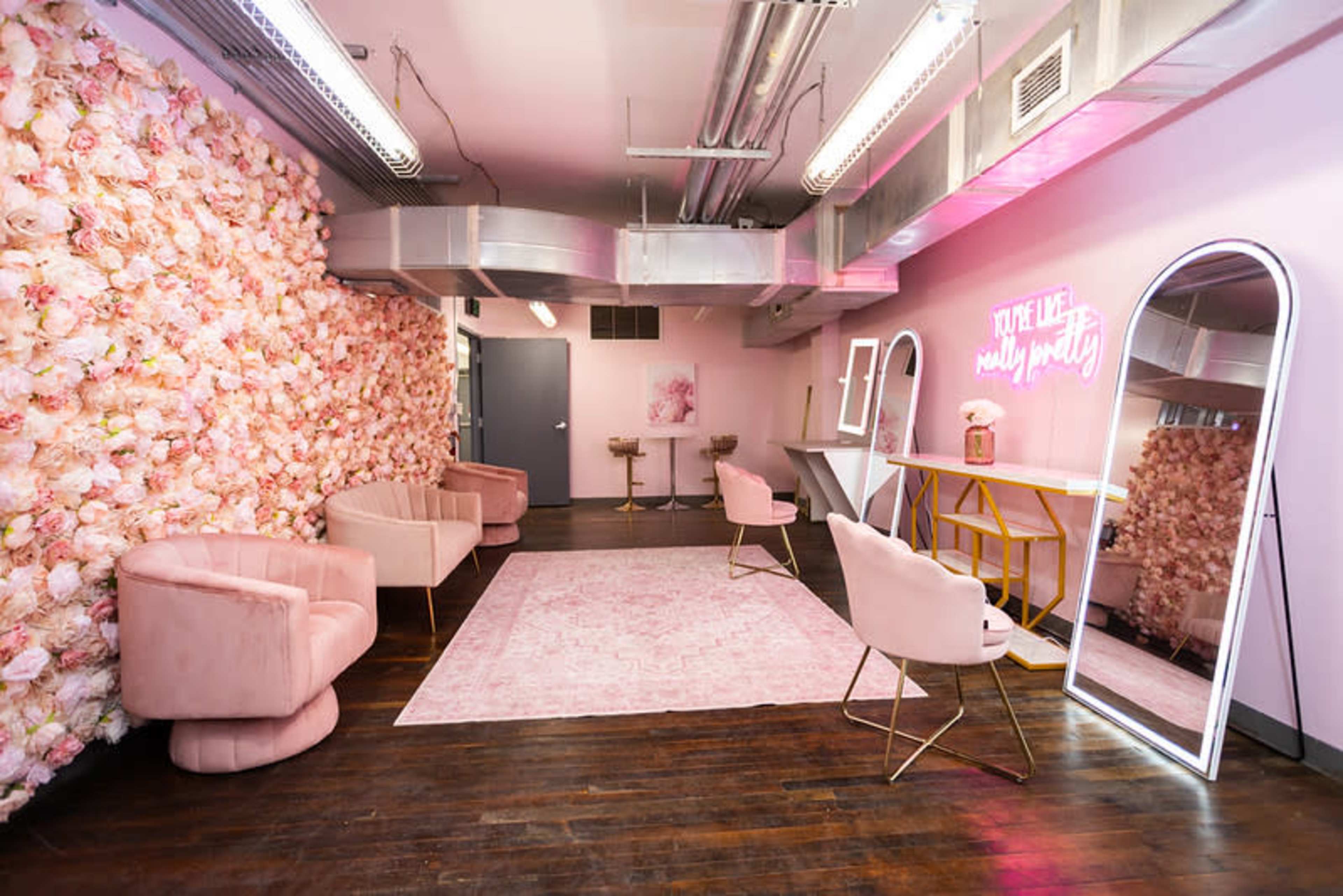 The image shows a softly lit room with a floral wall, stylish seating, and a vanity area featuring a neon sign.