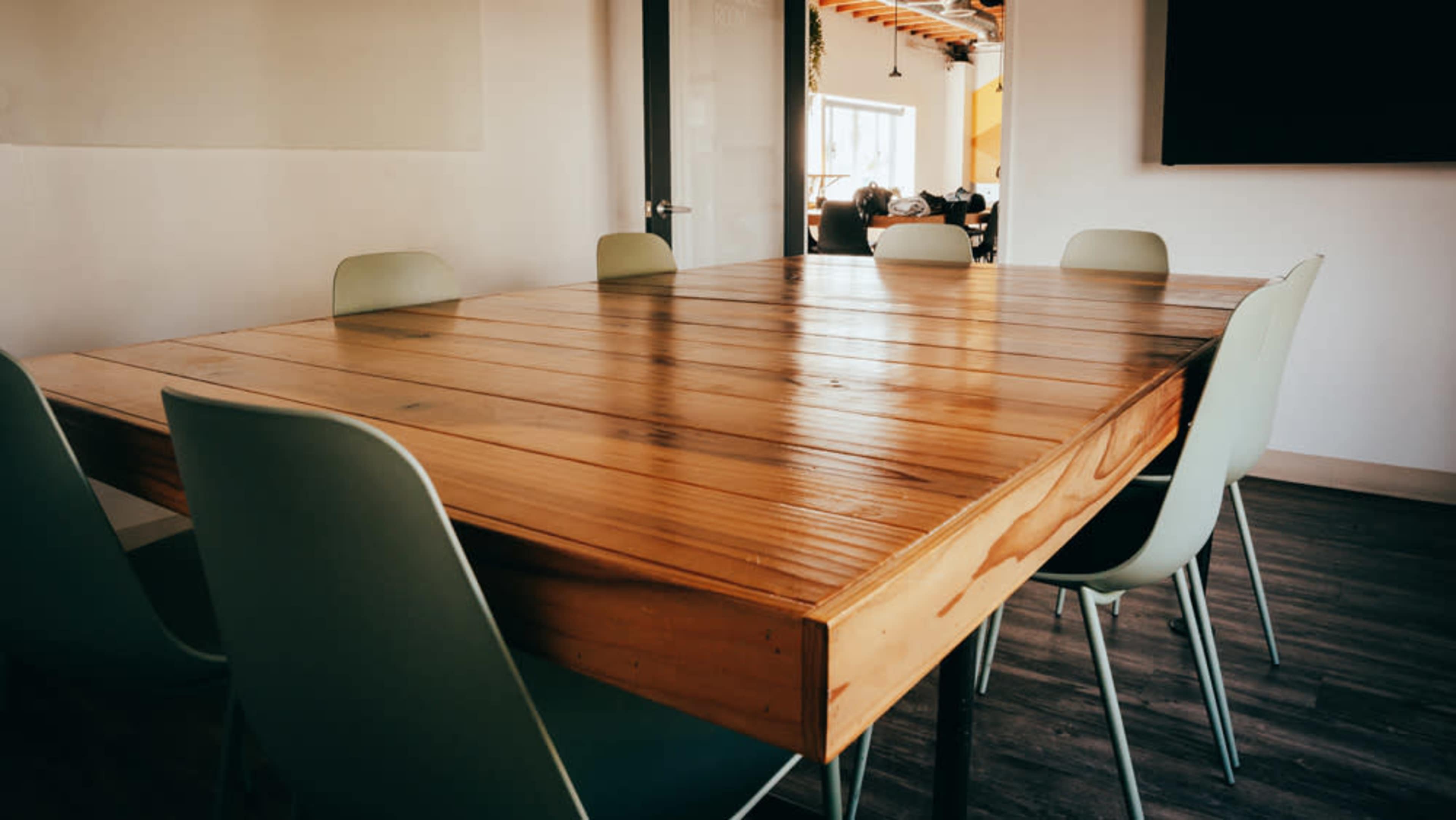 A large wooden table with a polished surface is surrounded by six light green chairs in a well-lit room.
