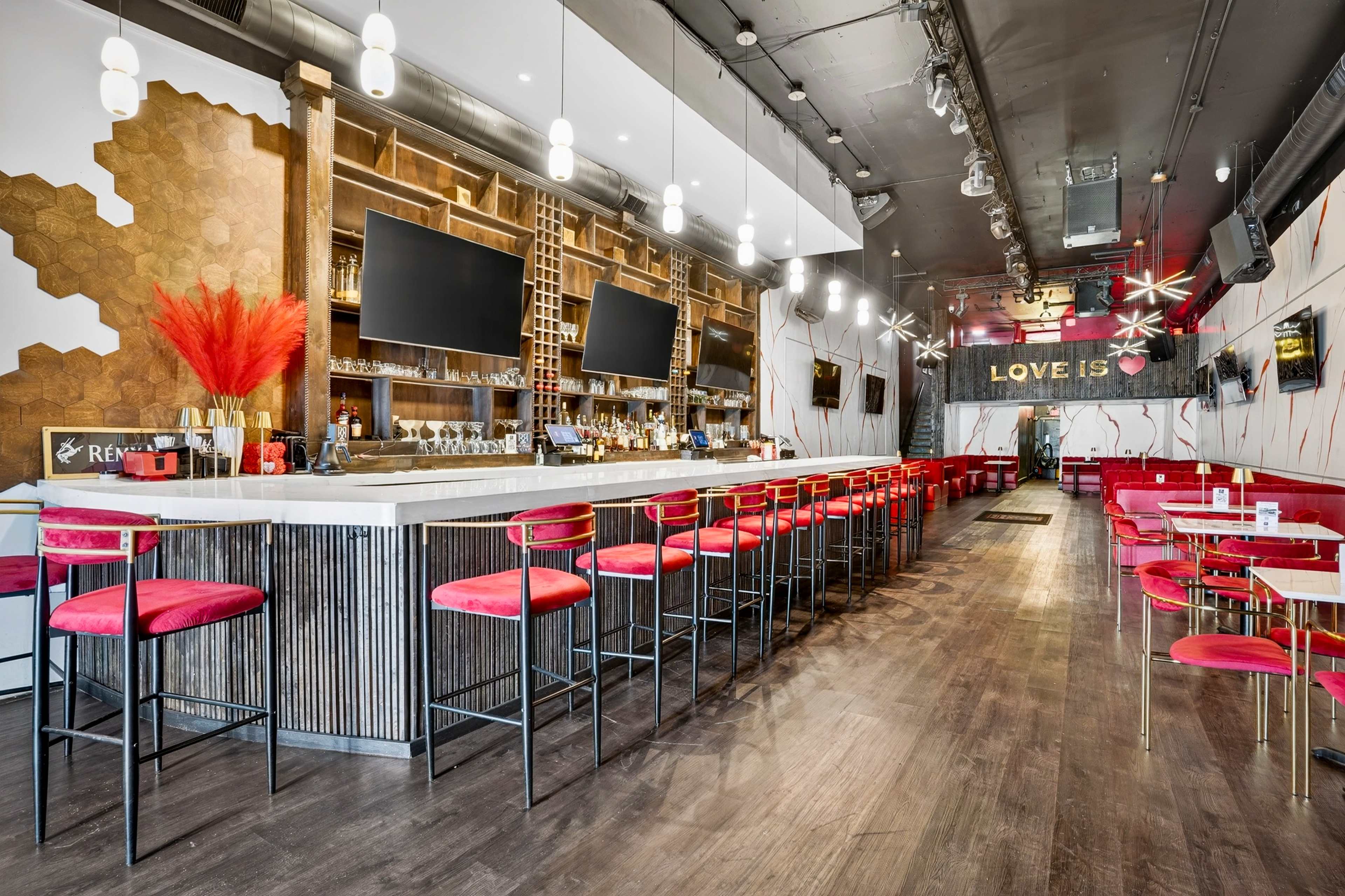 The image shows a modern bar with a long countertop, red bar stools, and large screens mounted on the wall.