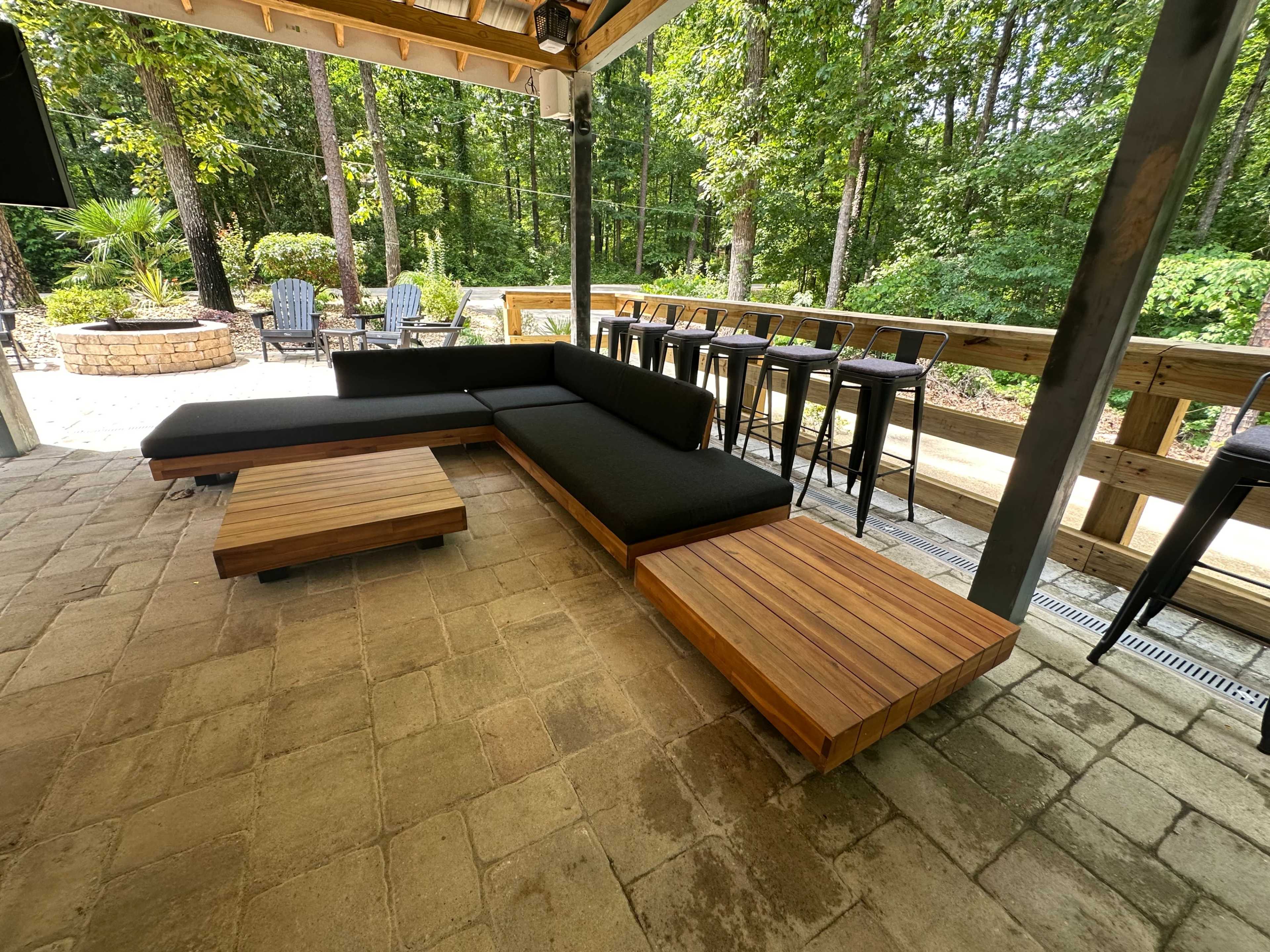 A modern outdoor seating area featuring a black sectional sofa with wooden coffee tables on a paved patio surrounded by trees and bar stools nearby.