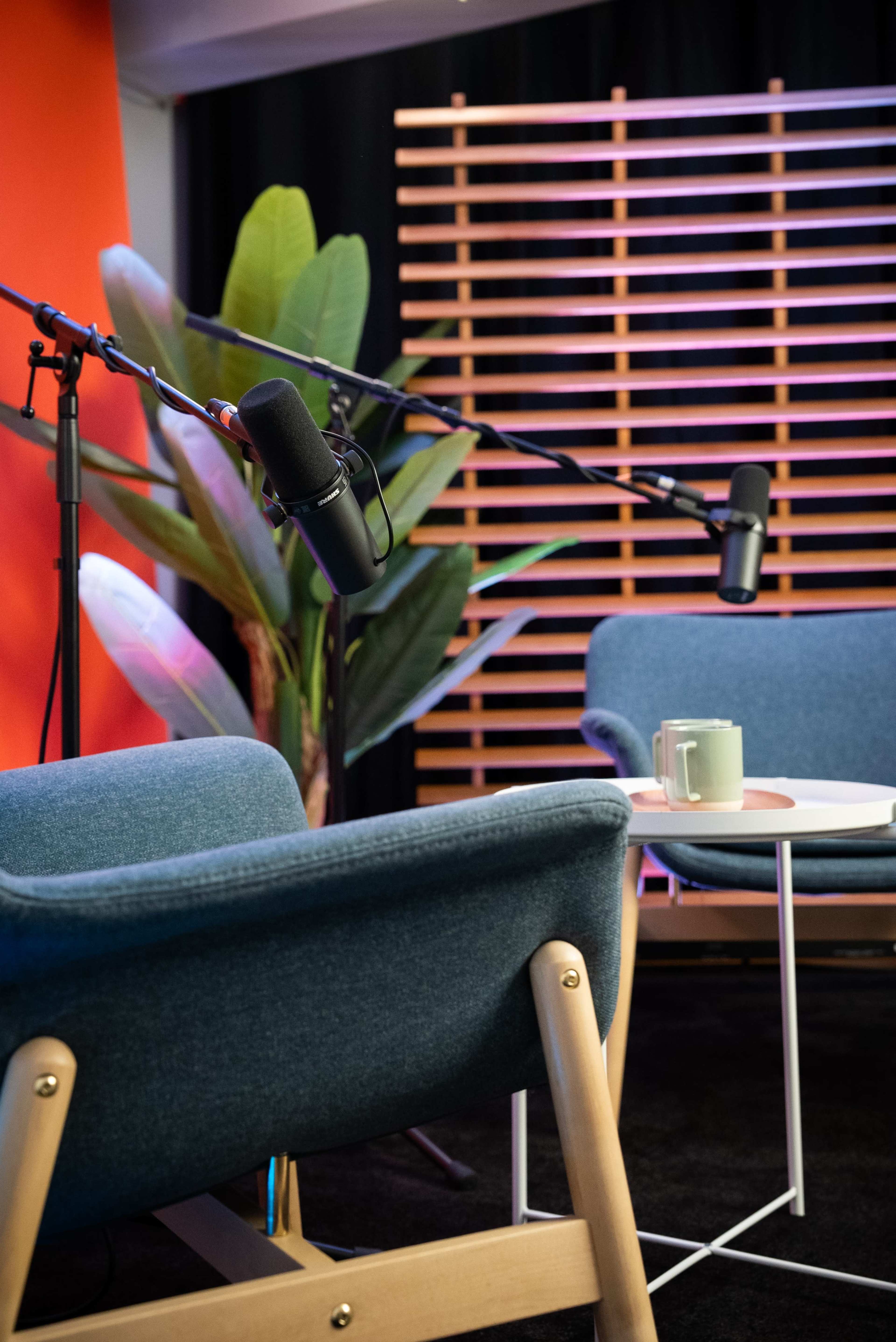 The image shows a modern interview setup with two blue chairs, a small white table, and microphones arranged in a studio-like environment.
