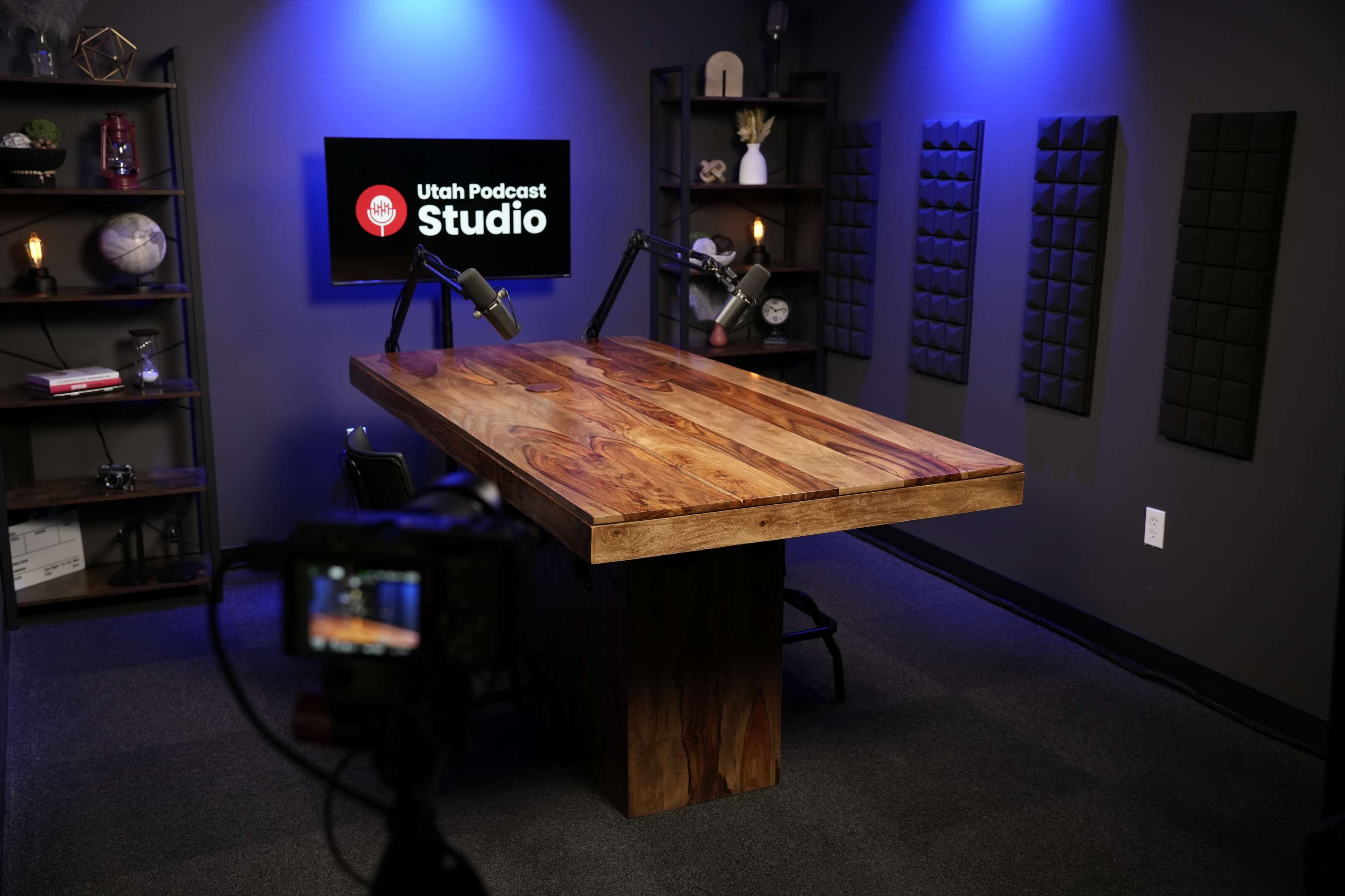 The image shows a podcast studio featuring a large wooden table, two microphones mounted on black stands, and a wall-mounted screen displaying the text "Utah Podcast Studio."