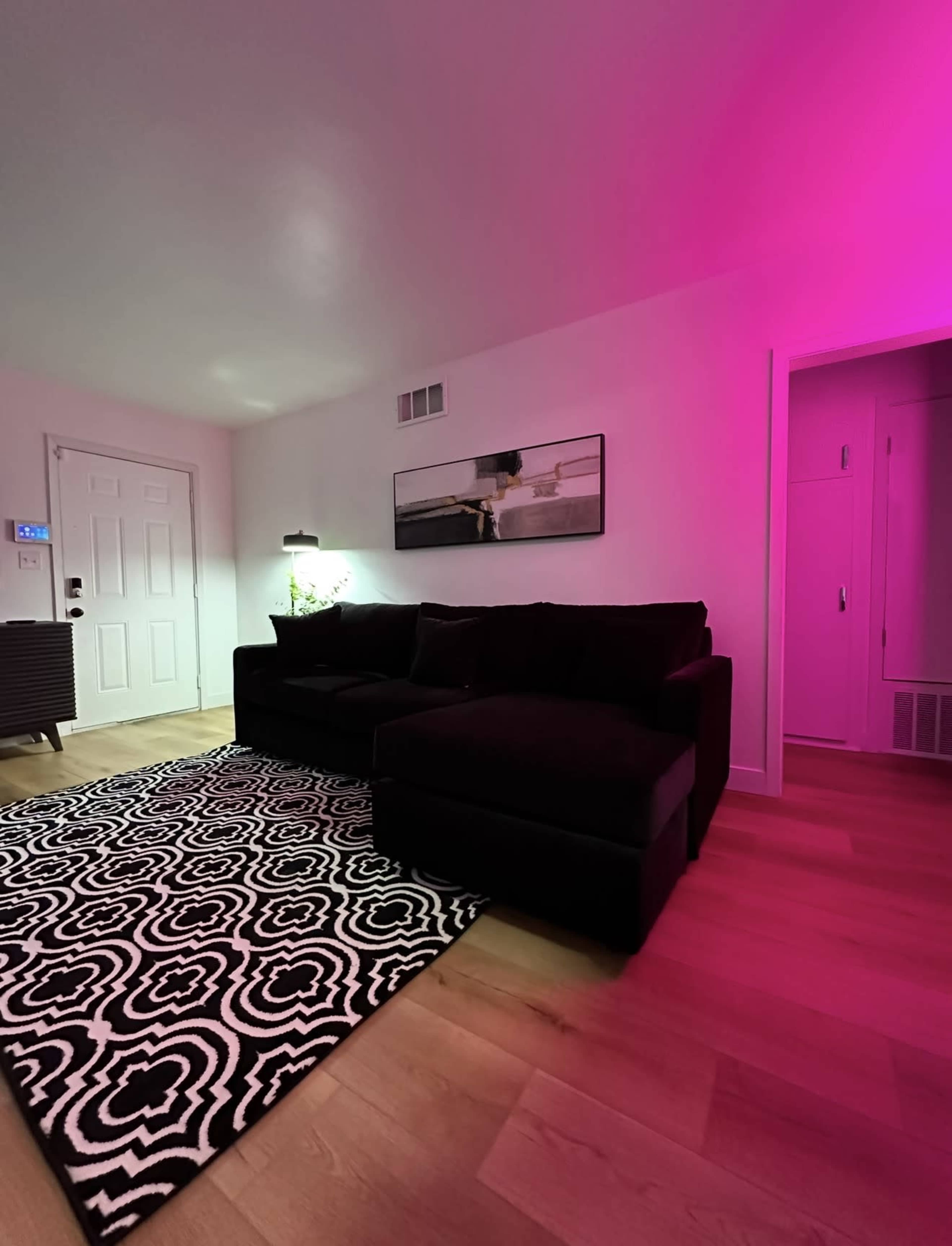The image shows a living room with a black couch, a patterned rug, and pink lighting along the walls.
