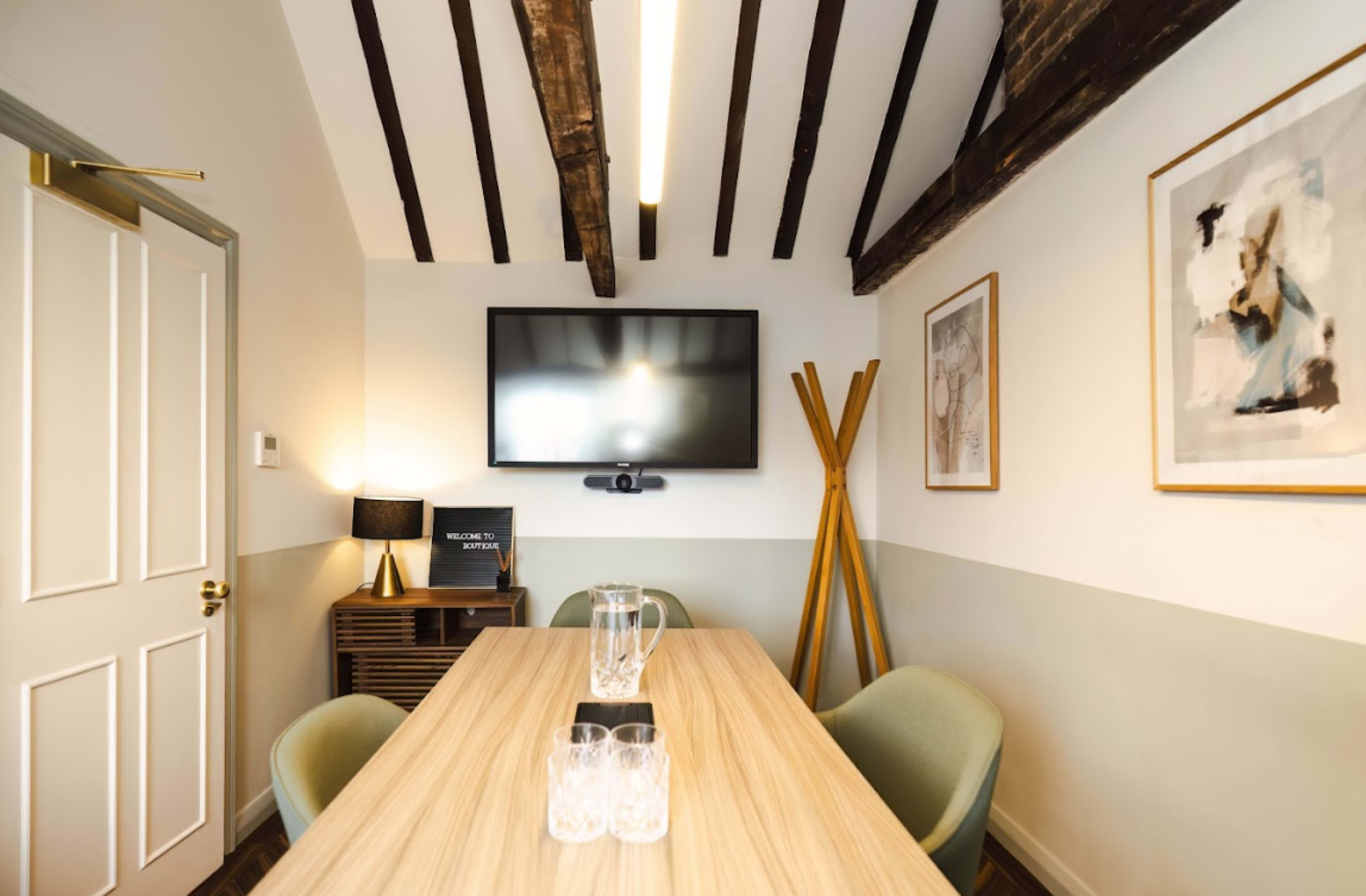 A small meeting room features a wooden table surrounded by stylish chairs, with a television mounted on the wall and decorative art pieces.