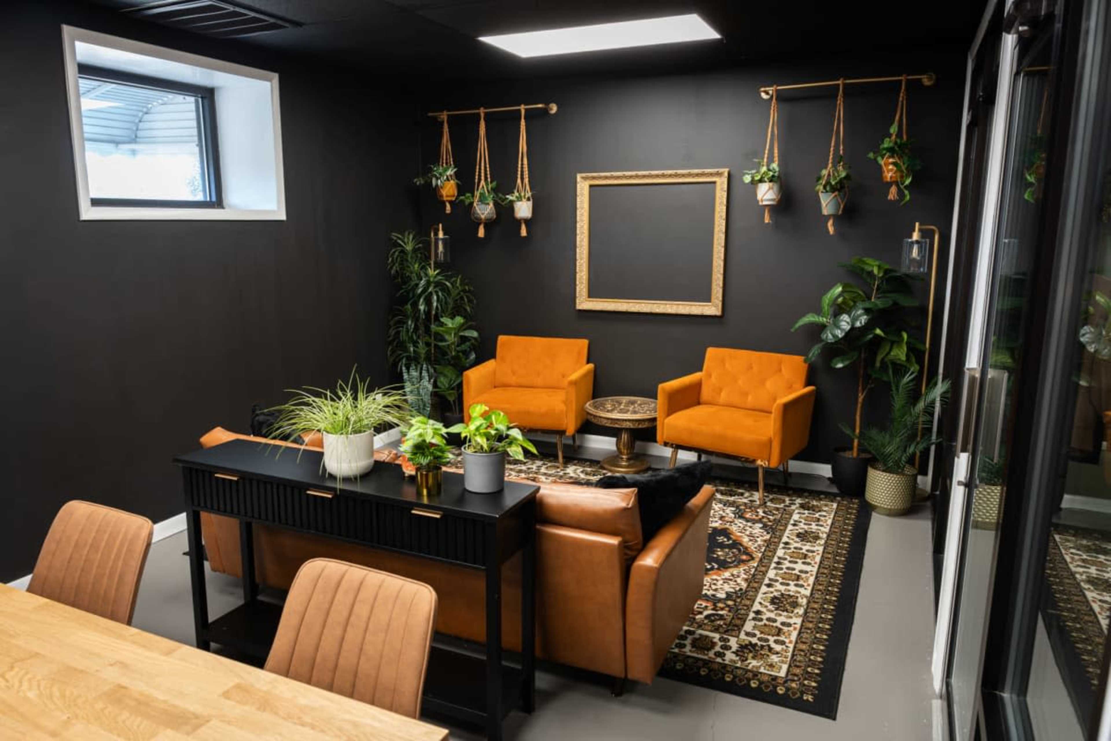 The image shows a modern indoor lounge area featuring two orange armchairs, a wooden table with plants, and various hanging planters on a dark wall.