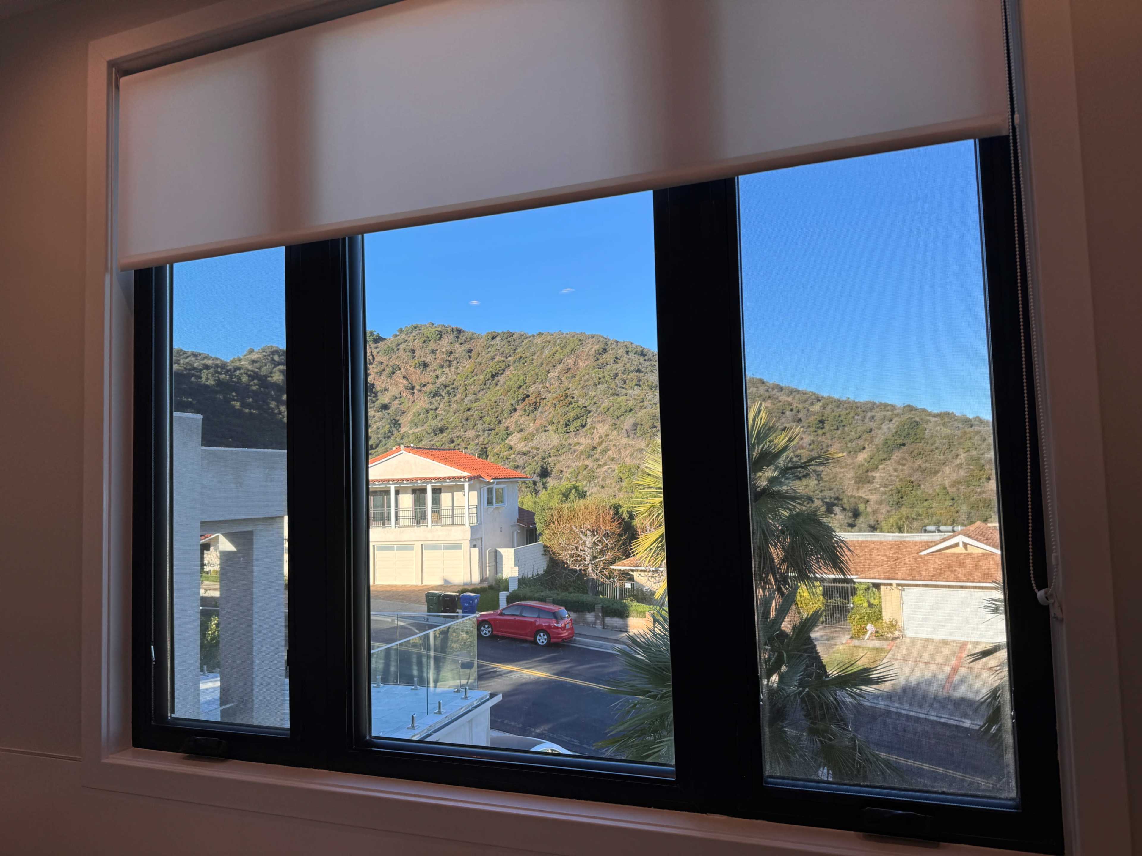 A window view overlooking a neighborhood with houses, a parked red car, and a hillside in the background.