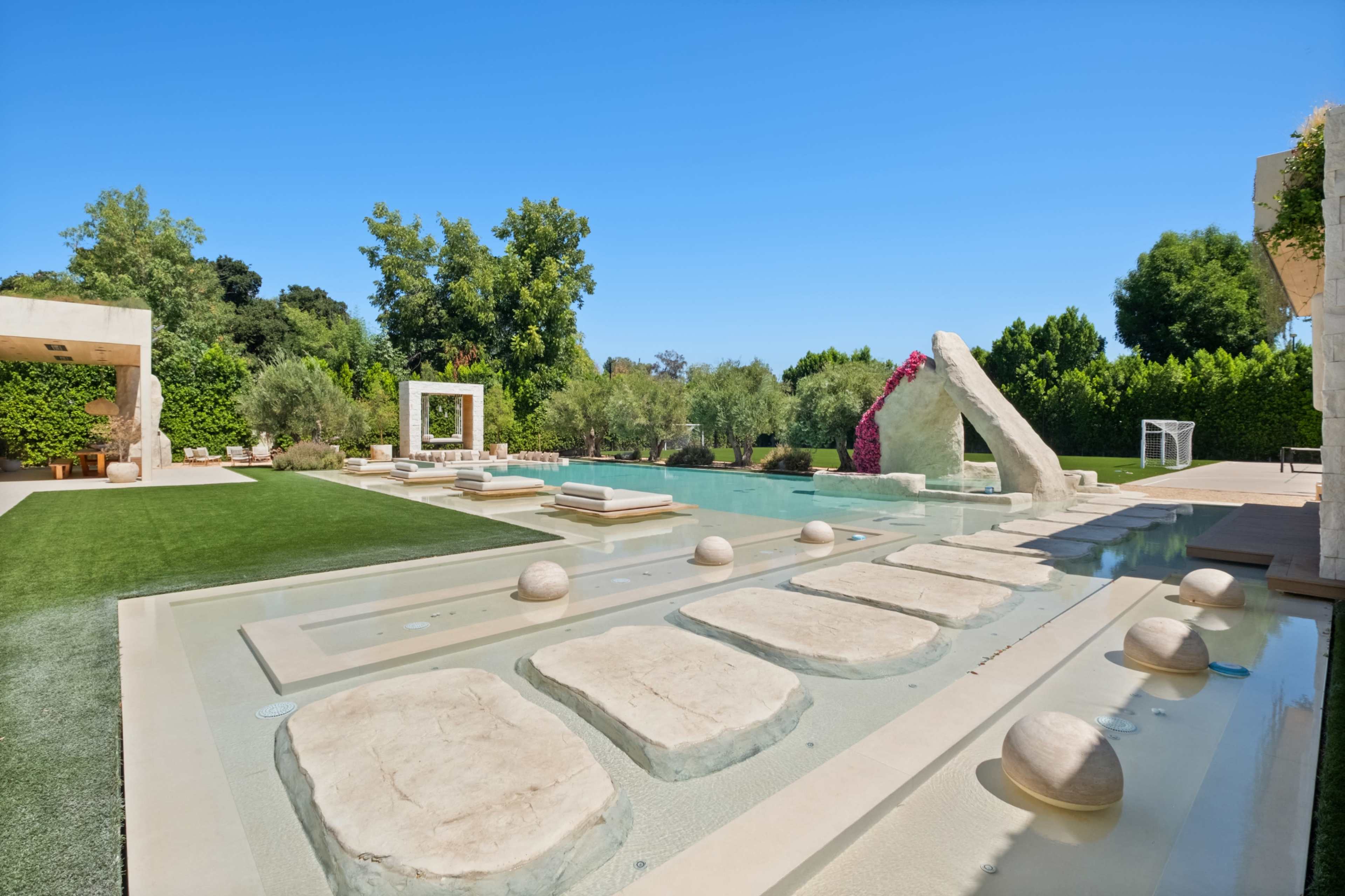 The image shows a modern outdoor pool area surrounded by landscaped gardens and stone pathways.