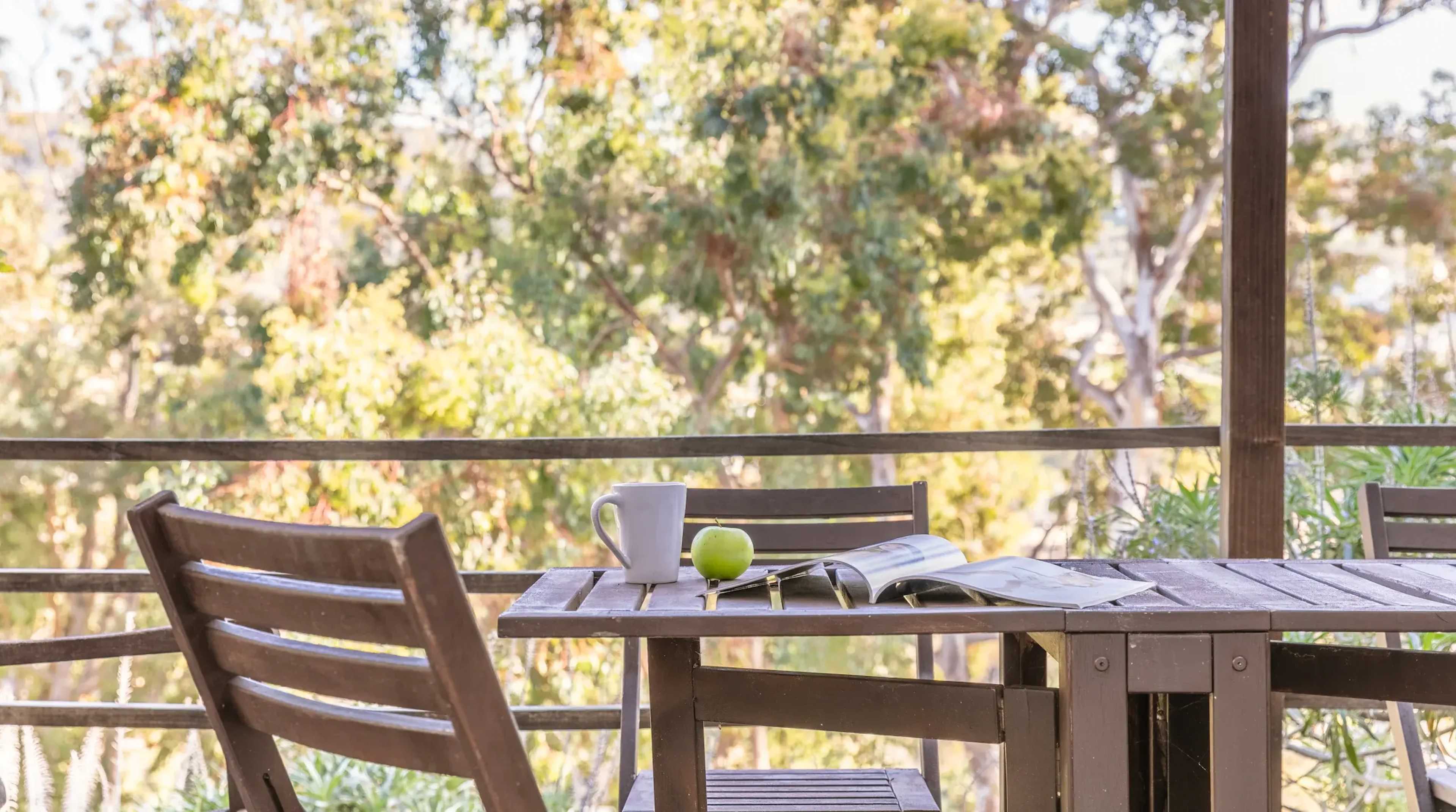 A wooden table with a magazine and a green apple sits on a deck overlooking trees in the background.
