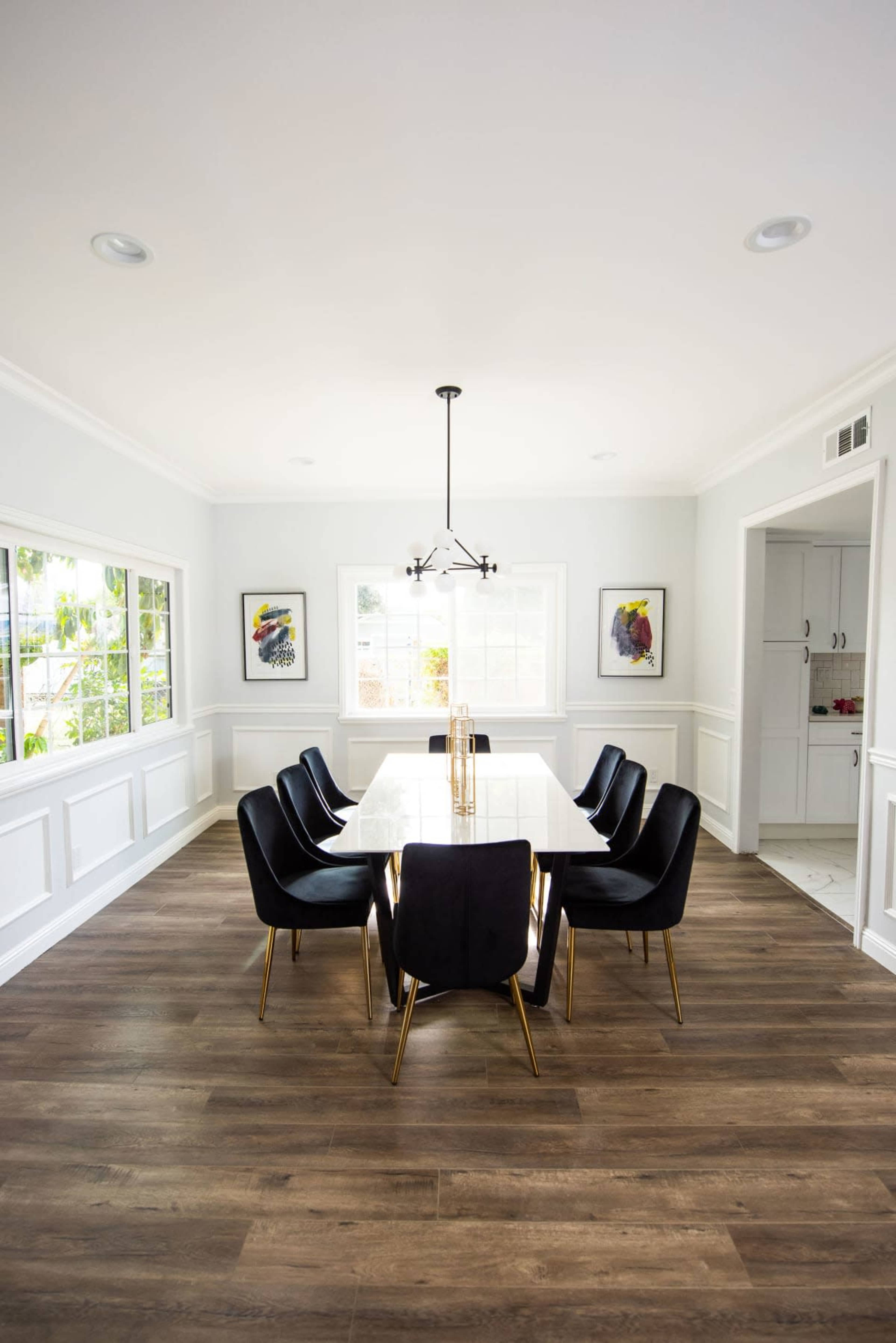 A modern dining room features a long glass table surrounded by six black chairs with gold legs, illuminated by a pendant light.