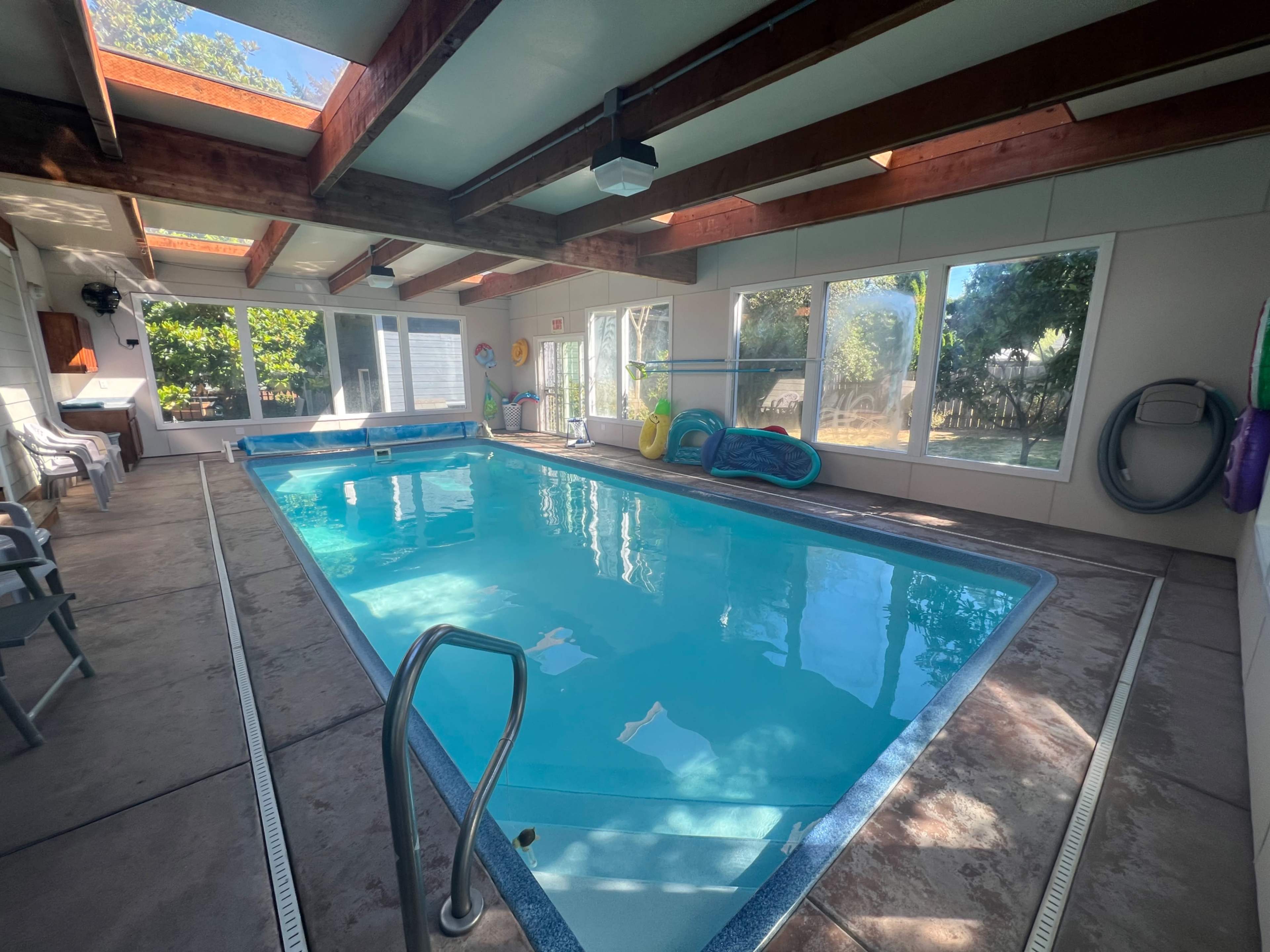 An indoor swimming pool surrounded by large windows, with lounge chairs and pool accessories nearby.