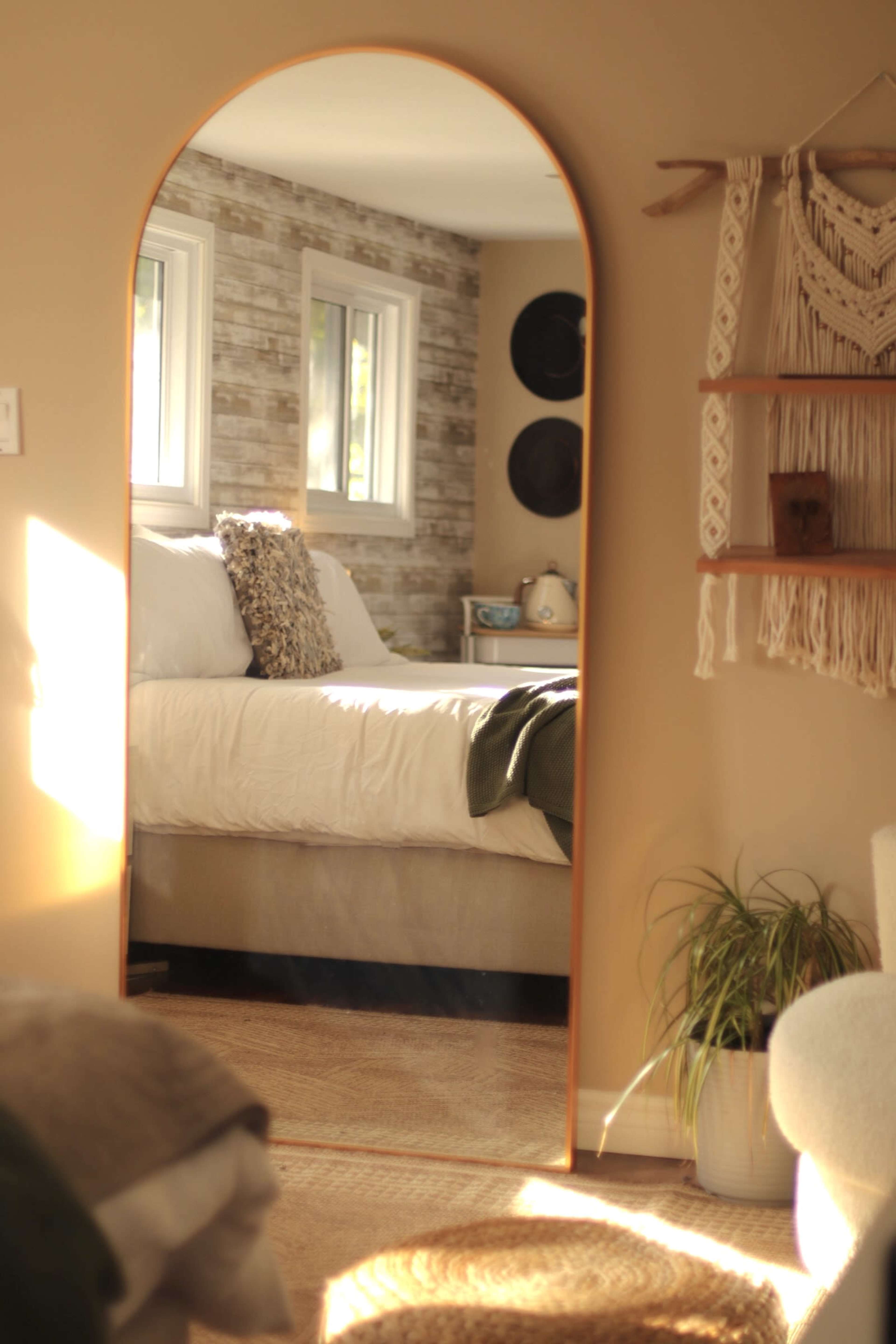 A large arched mirror reflects a cozy bedroom featuring a bed with white linens and a textured wall in the background.