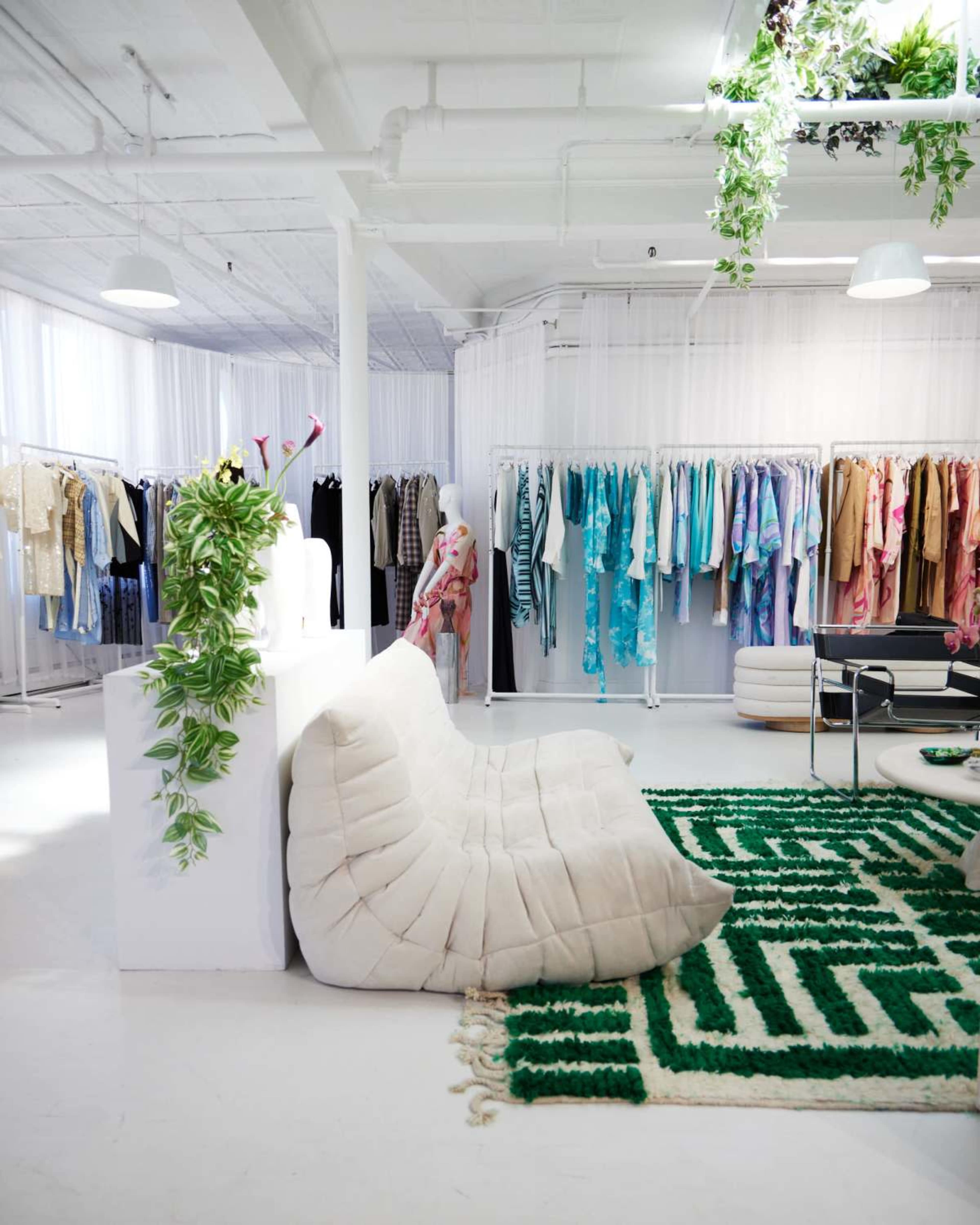 The image shows a modern clothing showroom with colorful apparel displayed on racks, a plush white seating area, and a green patterned rug.