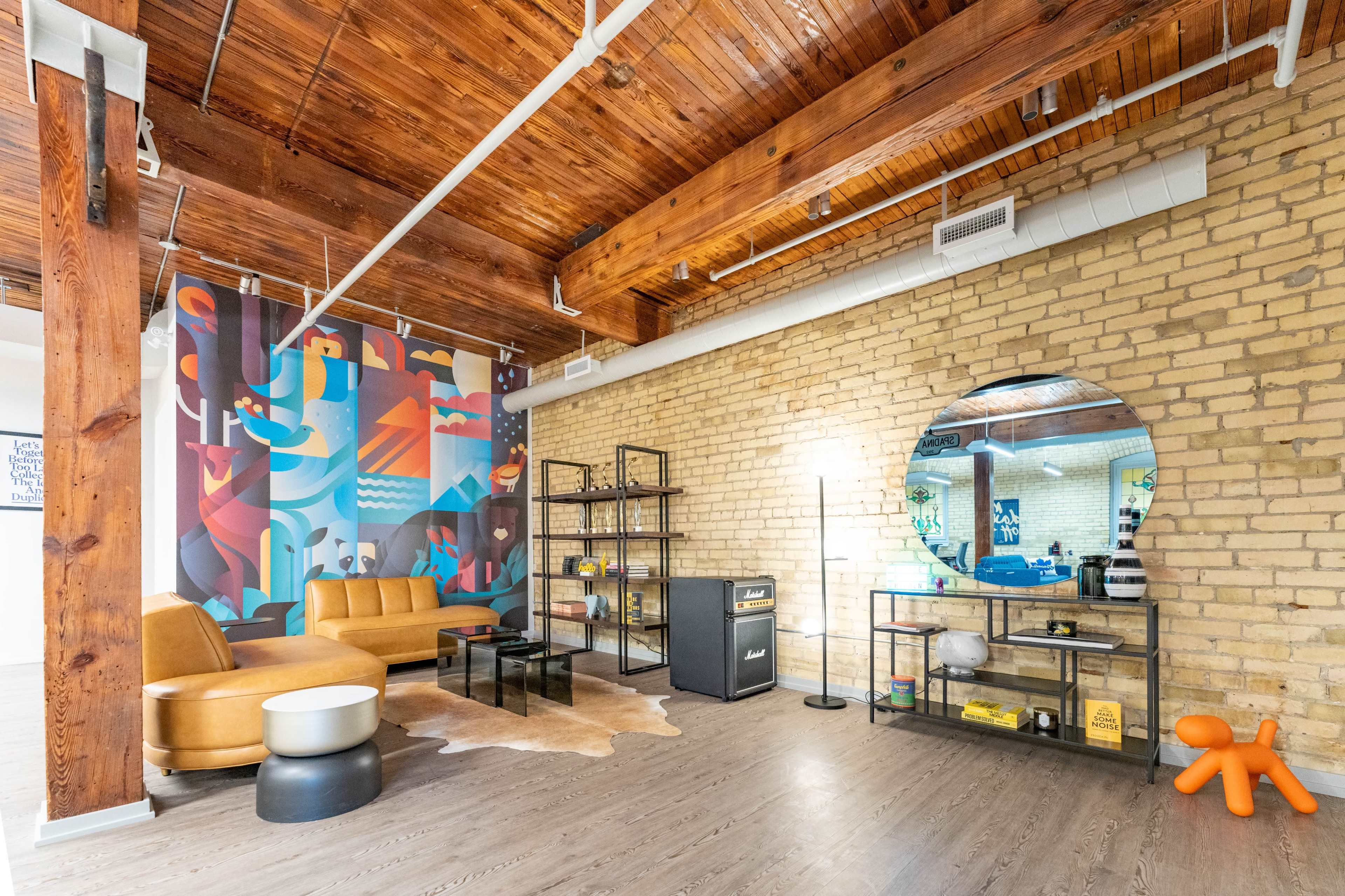A modern lounge area with a brown sectional sofa, a round coffee table, a large mirror, and bookshelves against a brick wall with colorful artwork.