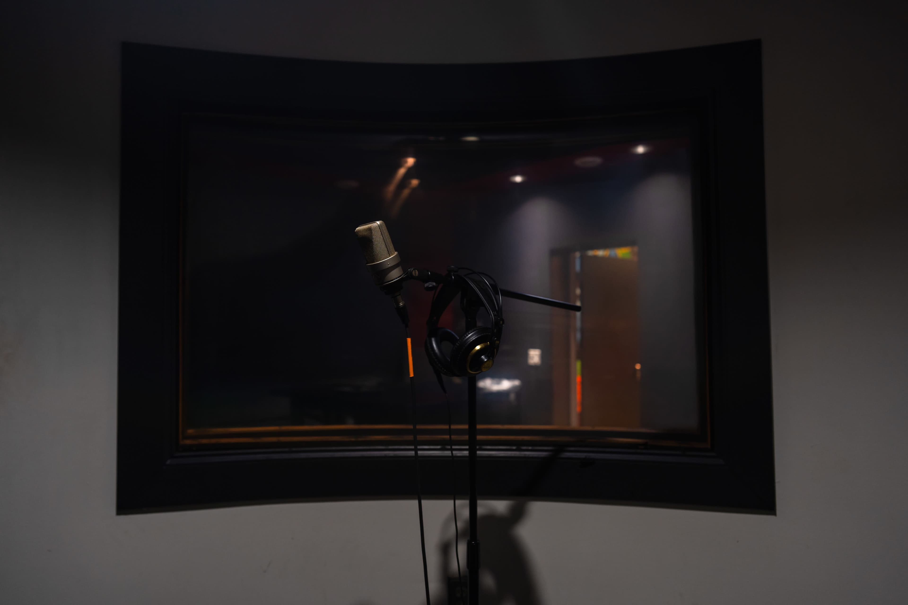 A microphone and headphones are positioned on a stand in a sound recording studio, with a glass window in the background.