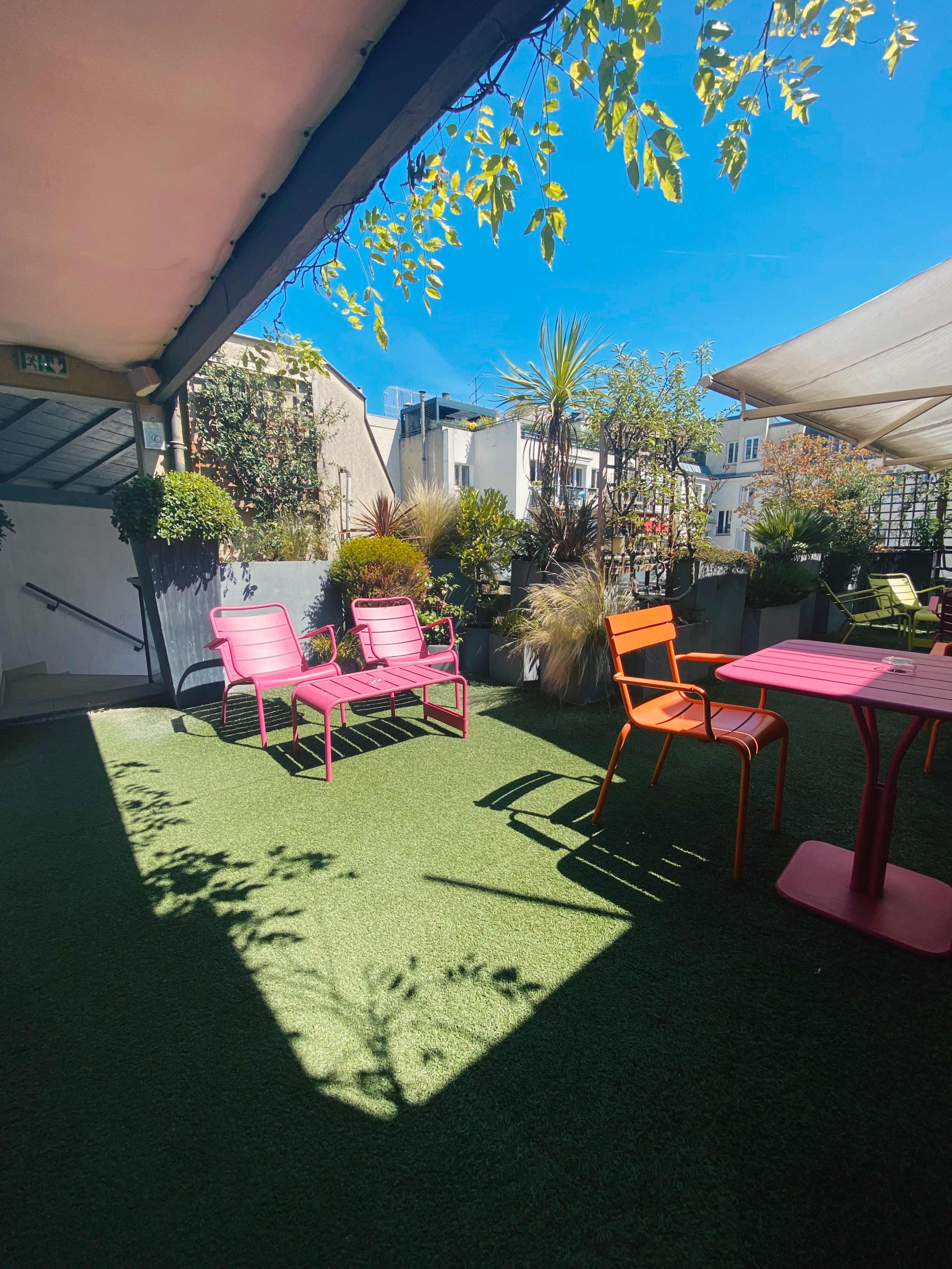 A rooftop terrace with pink chairs and a table surrounded by greenery under a clear blue sky.