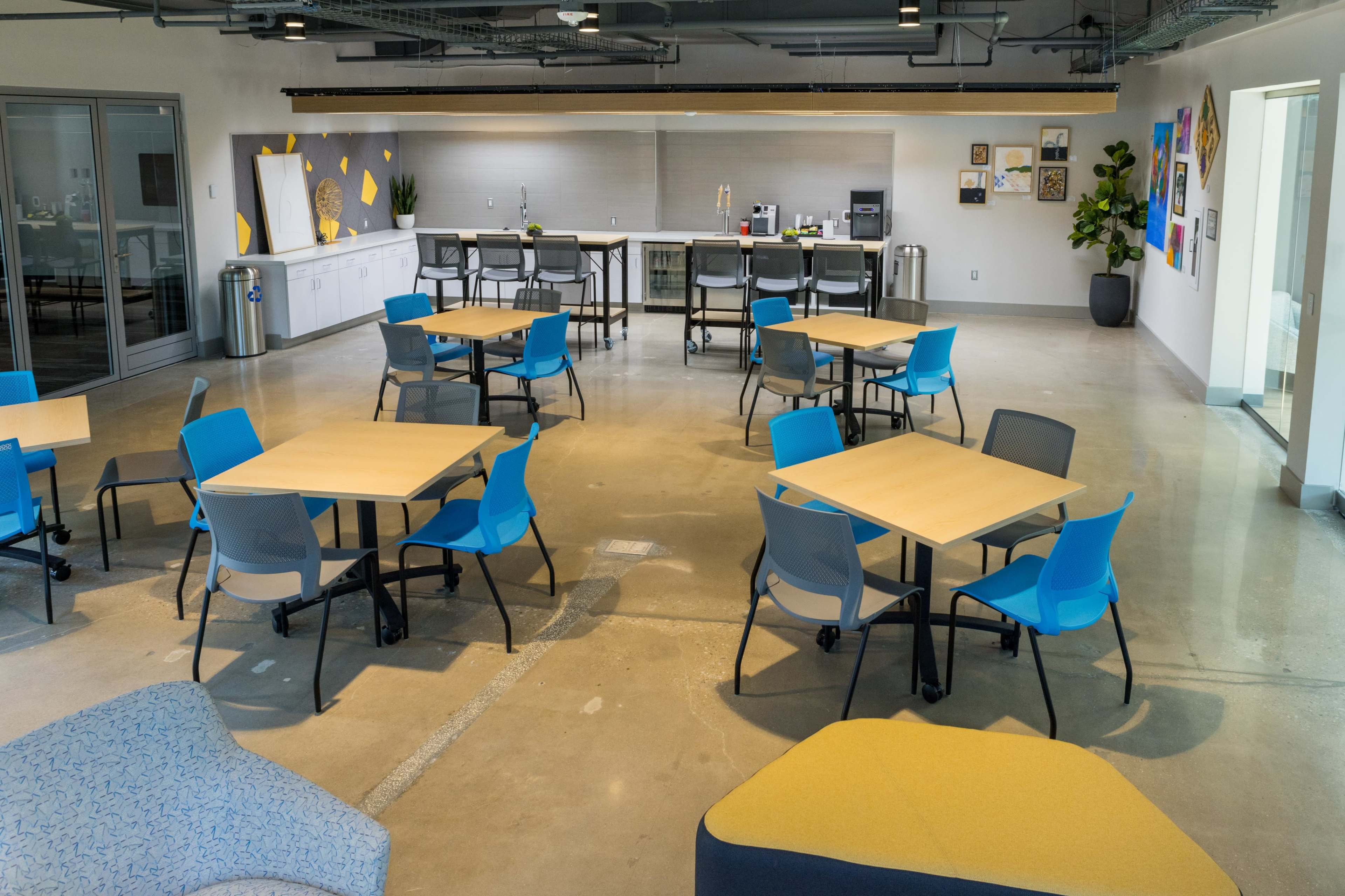 The image shows a modern office break room with tables and chairs arranged in a sparse layout, along with a kitchenette area and decorative wall art in the background.