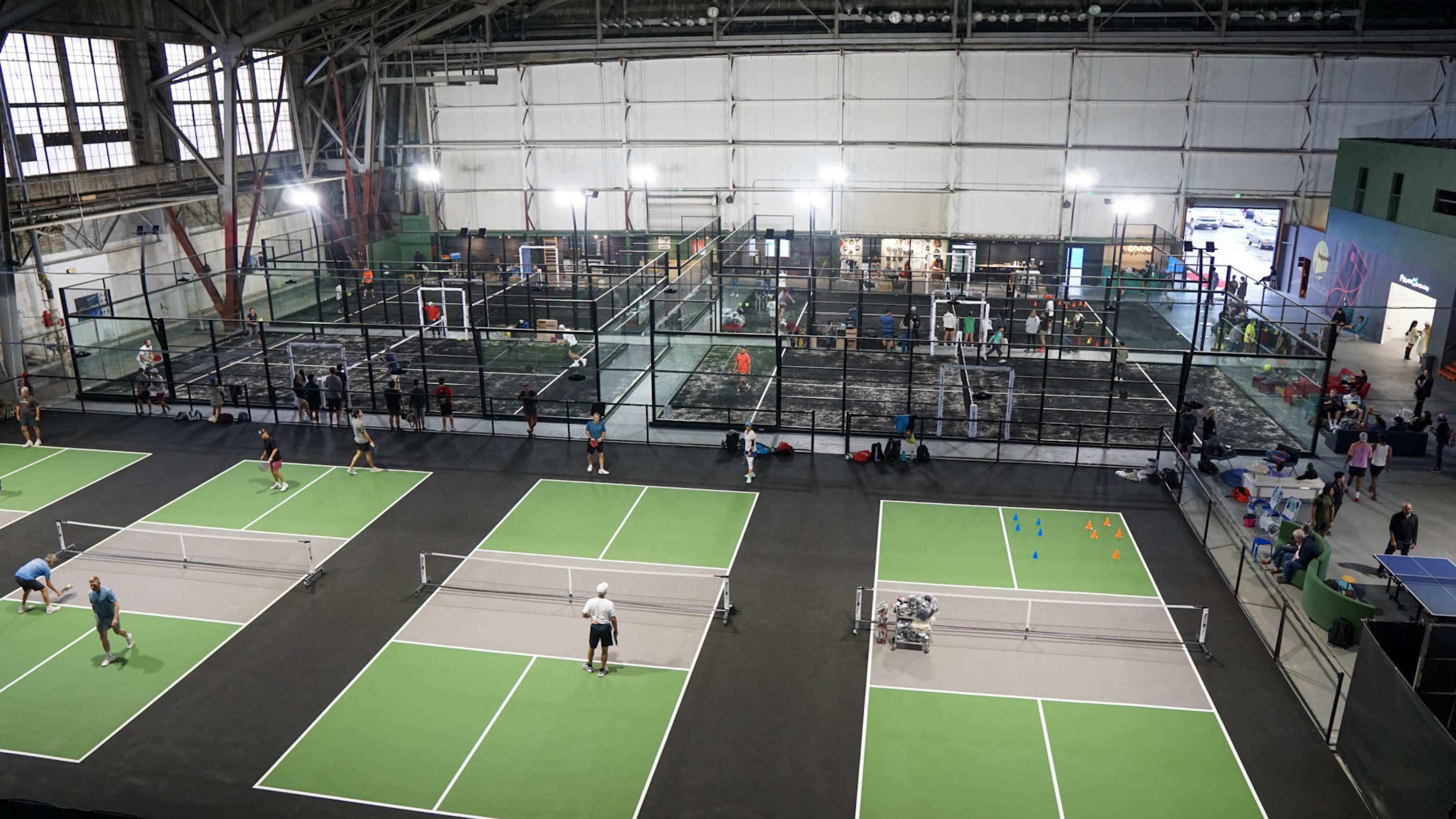 The image shows an indoor sports facility featuring multiple courts for tennis and pickleball, with players actively participating in various games.