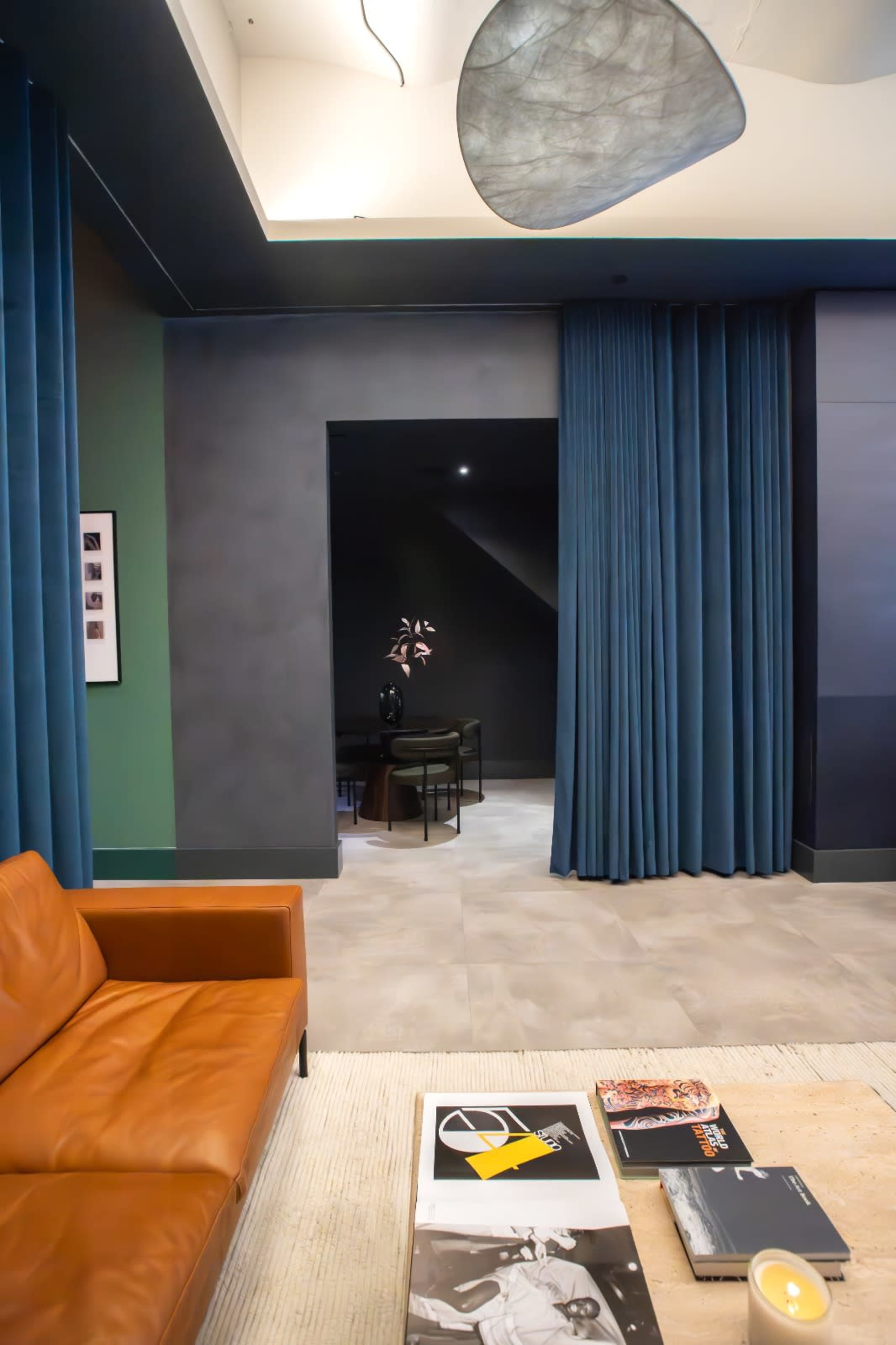 A contemporary living space features a brown leather sofa, a coffee table with magazines, and dark curtains framing a doorway leading to another room.