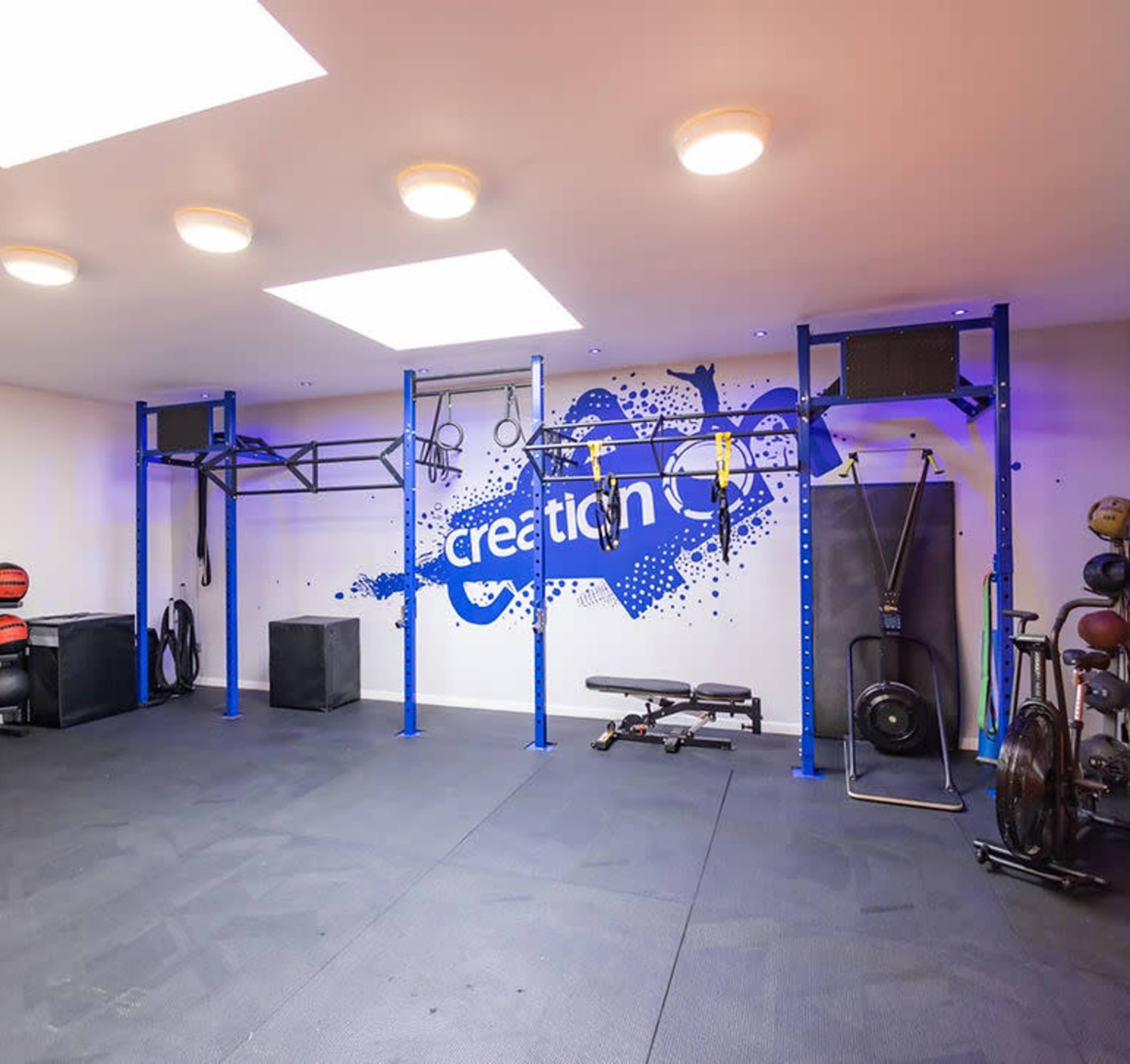 The image shows a well-equipped gym space featuring exercise equipment, a pull-up rig, and a blue wall with a graphic design.