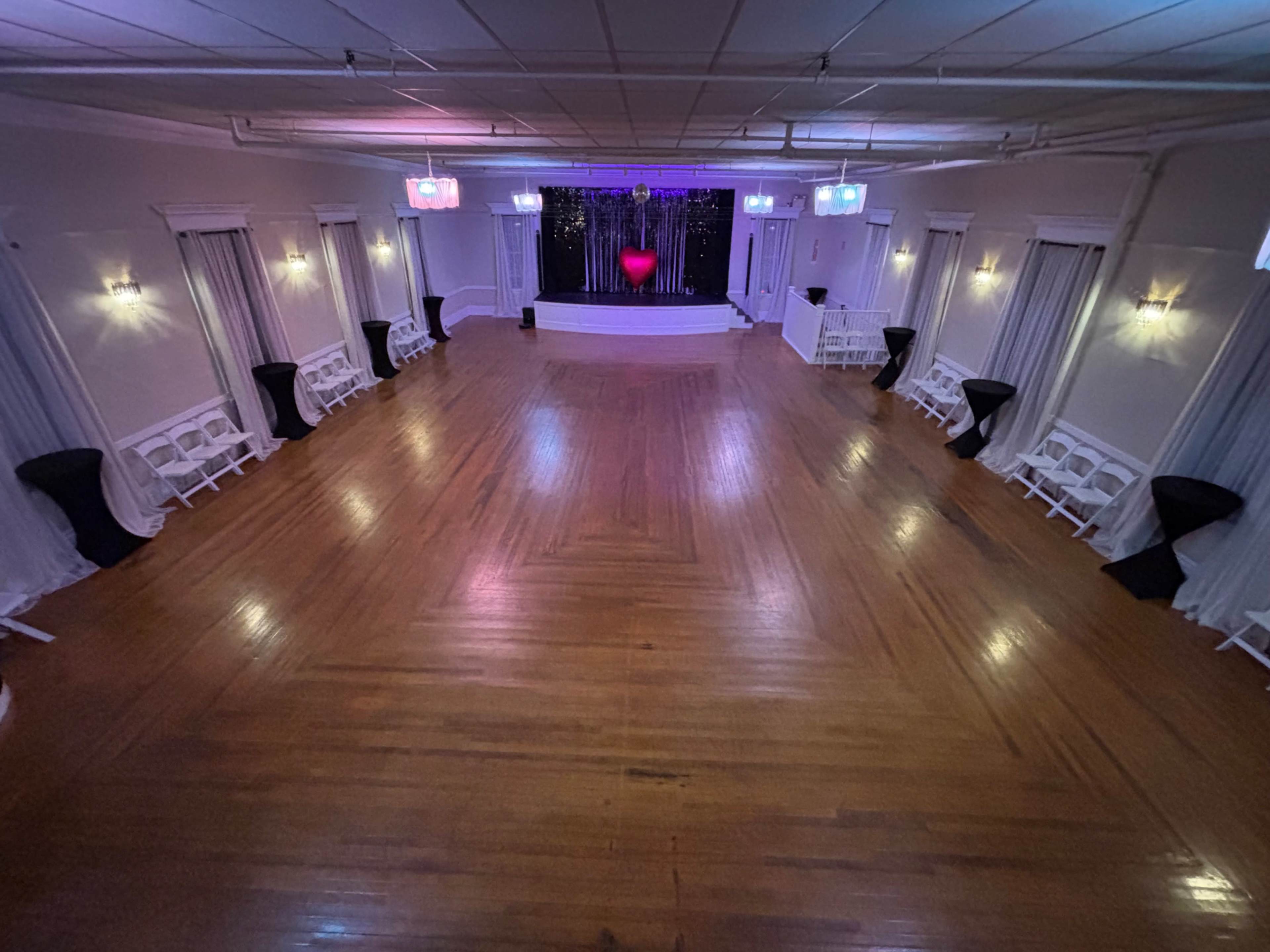The image shows an empty event space with polished wooden floors, draped curtains, and a stage at the front adorned with colored lights.
