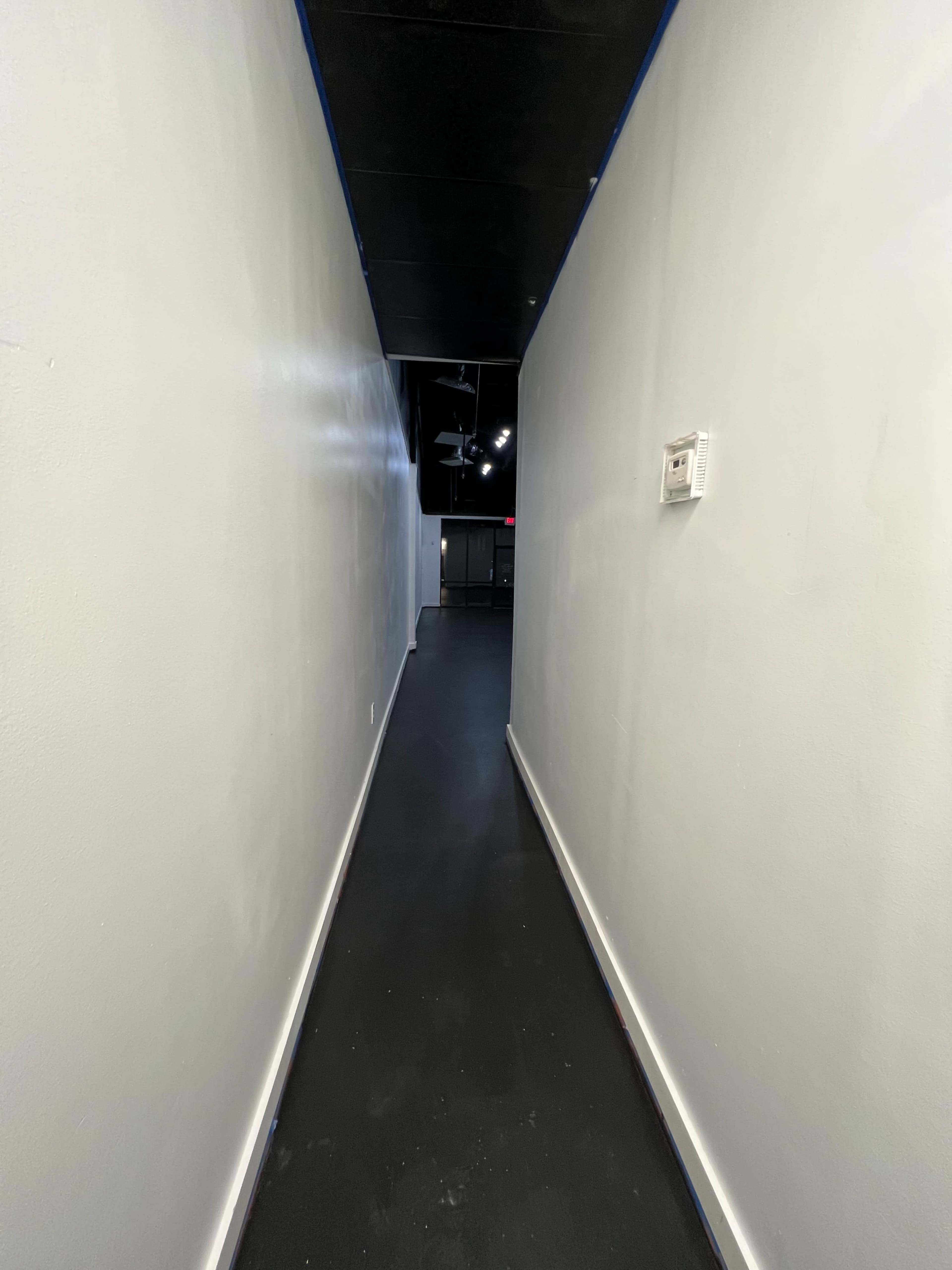 The image shows a narrow, dimly lit hallway with white walls and a black floor leading to a doorway at the end.