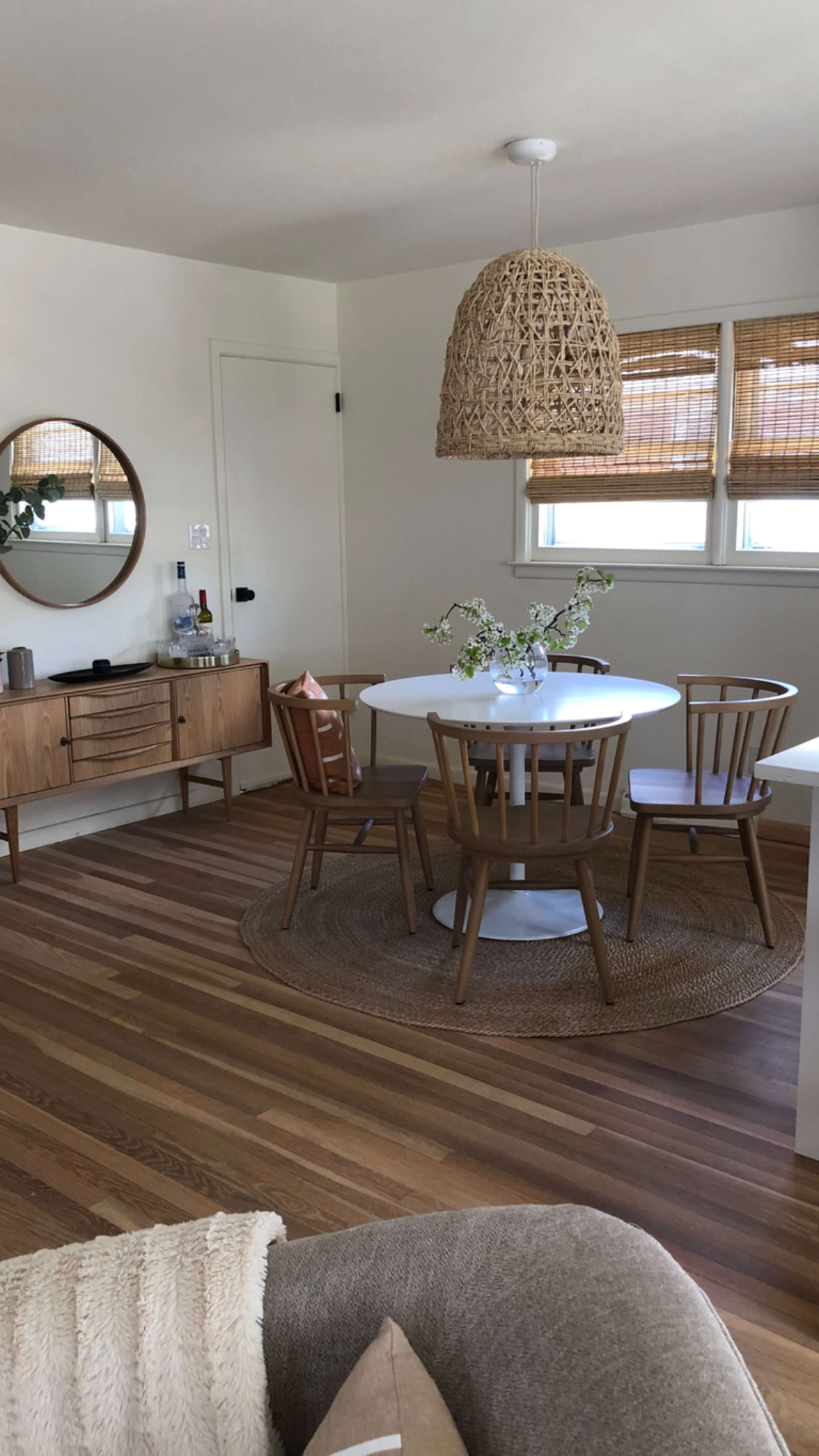 A round white dining table surrounded by wooden chairs is positioned in a light, spacious room with a woven pendant light and a sideboard.