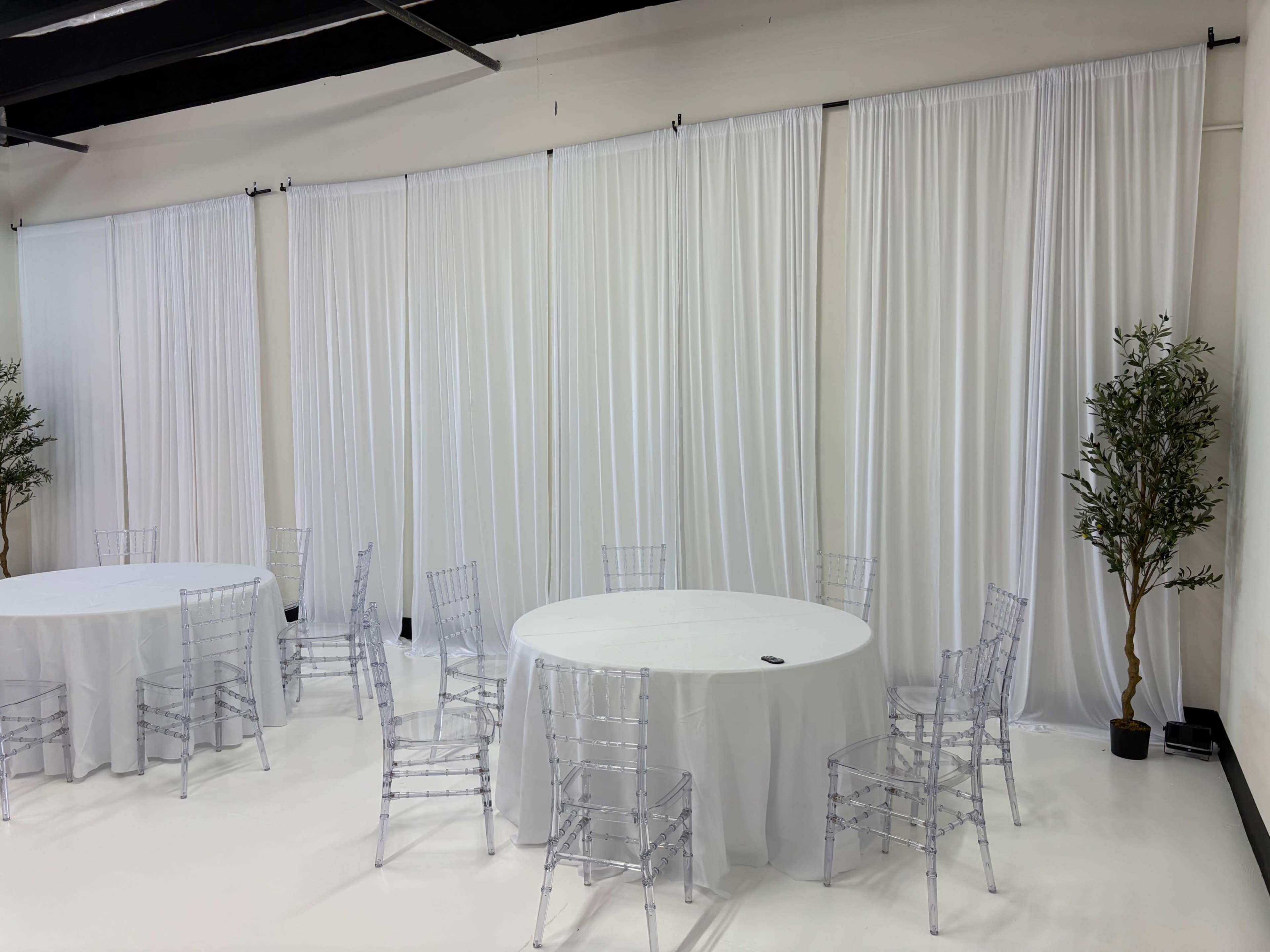The image shows a minimalist event space with three round tables covered in white tablecloths, surrounded by clear ghost chairs, and white drapes hanging on the walls.
