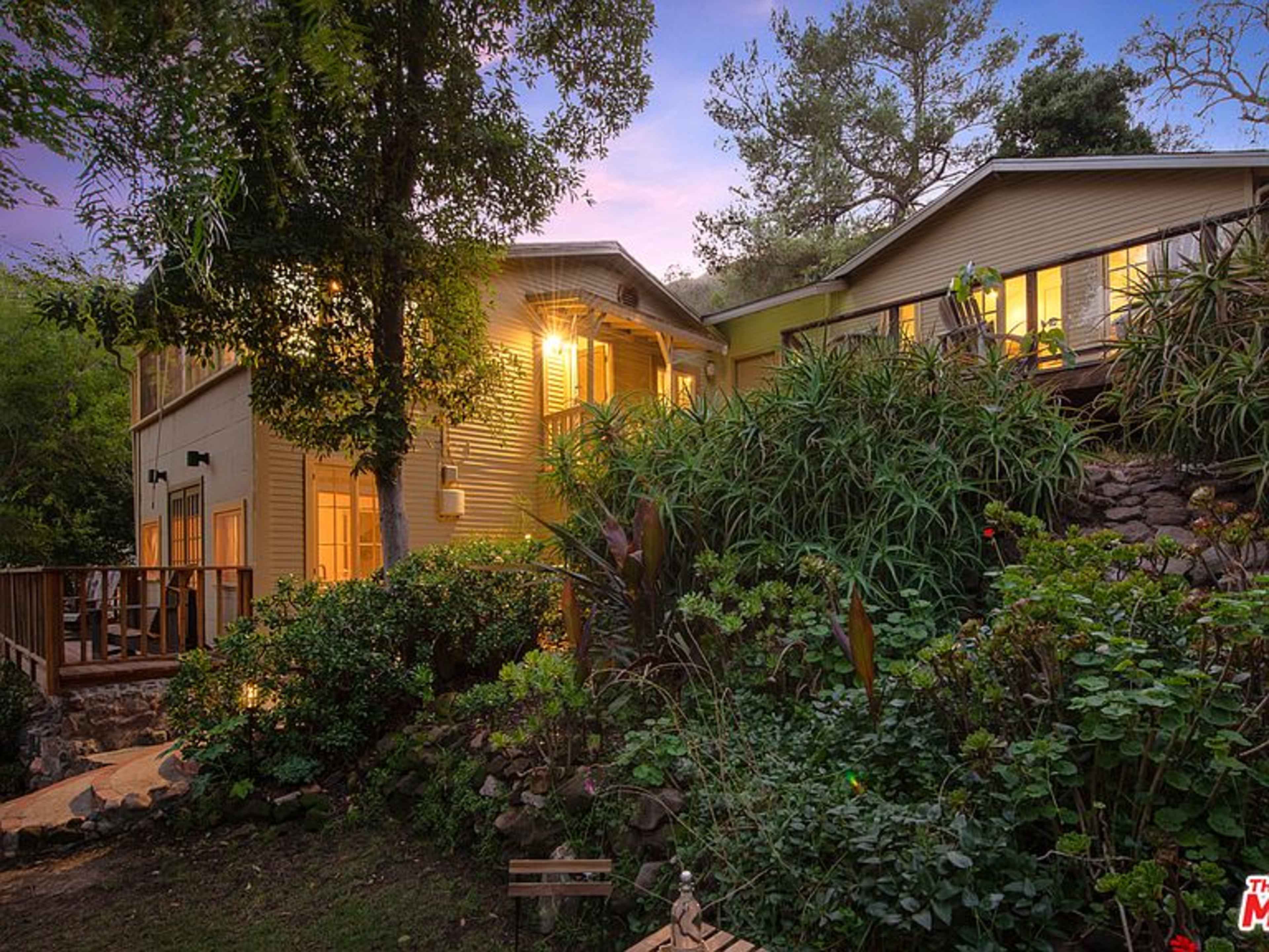 A two-story house surrounded by lush greenery and gardens at dusk, with warm lights illuminating the windows.