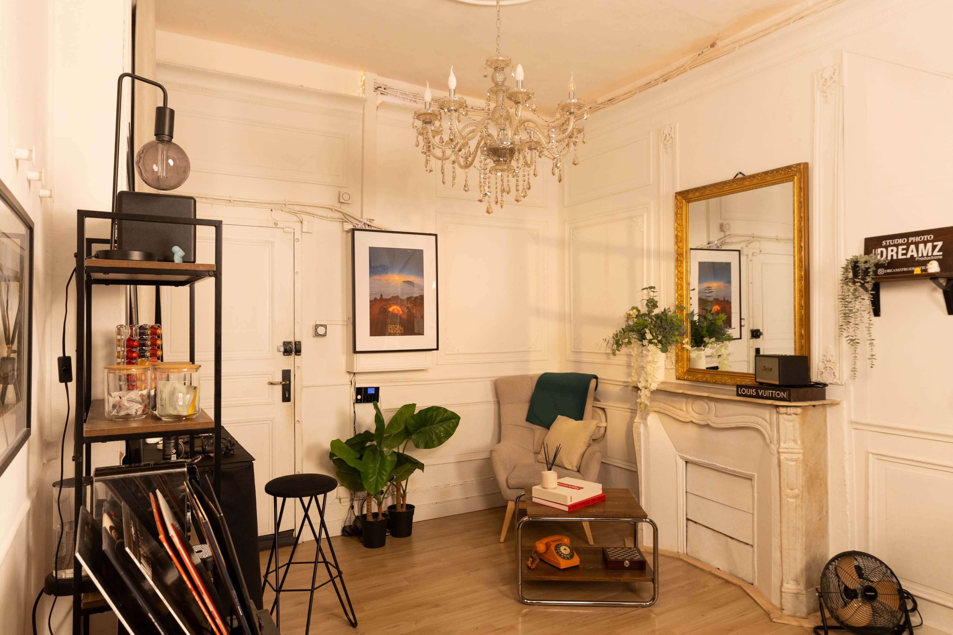 The image shows a well-decorated corner of a room featuring a chandelier, a large mirror, potted plants, and a mix of furniture including a stool and a chair.