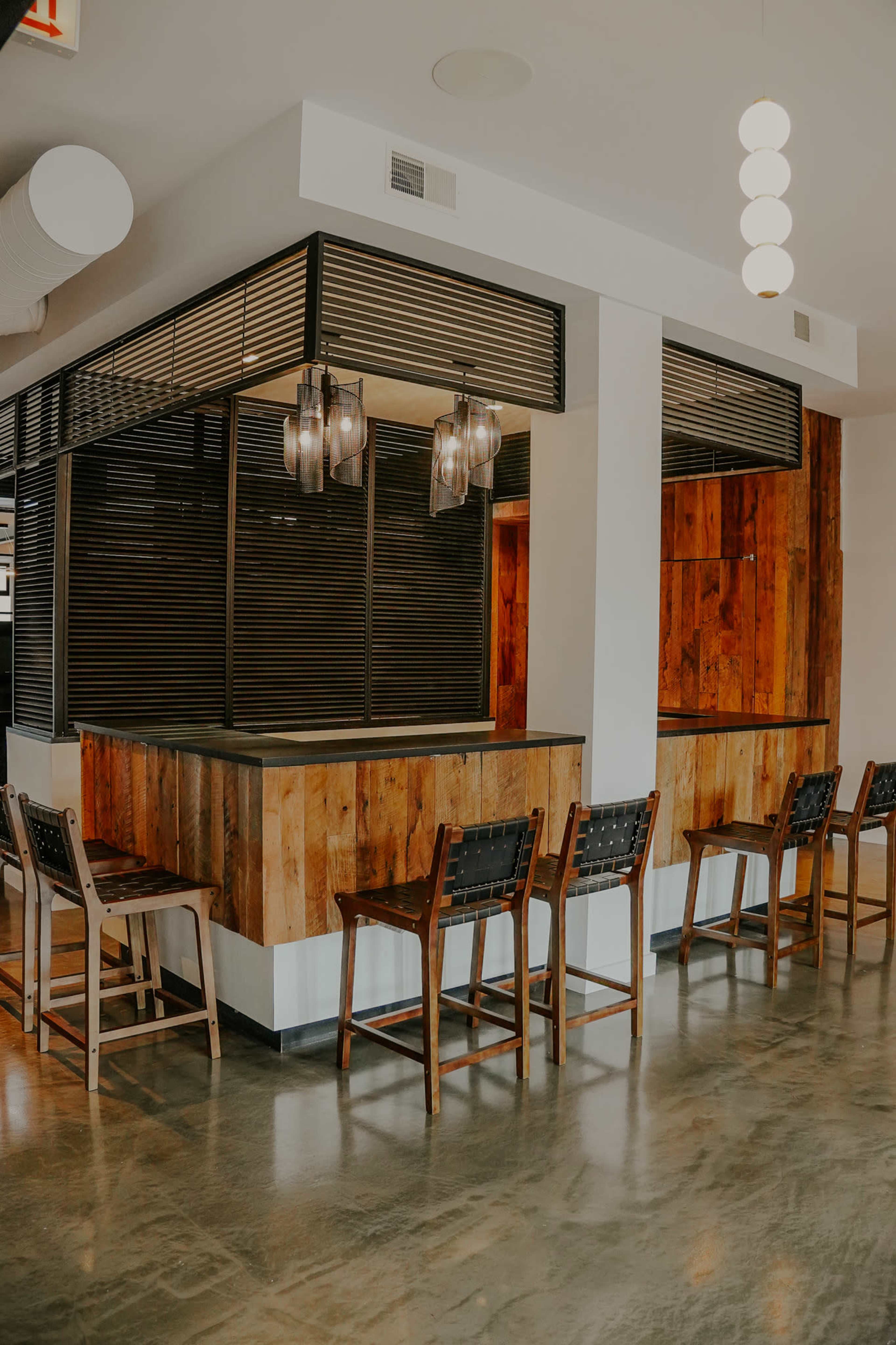 The image shows a modern interior of a bar area with wooden accents, high stools, and pendant lighting.