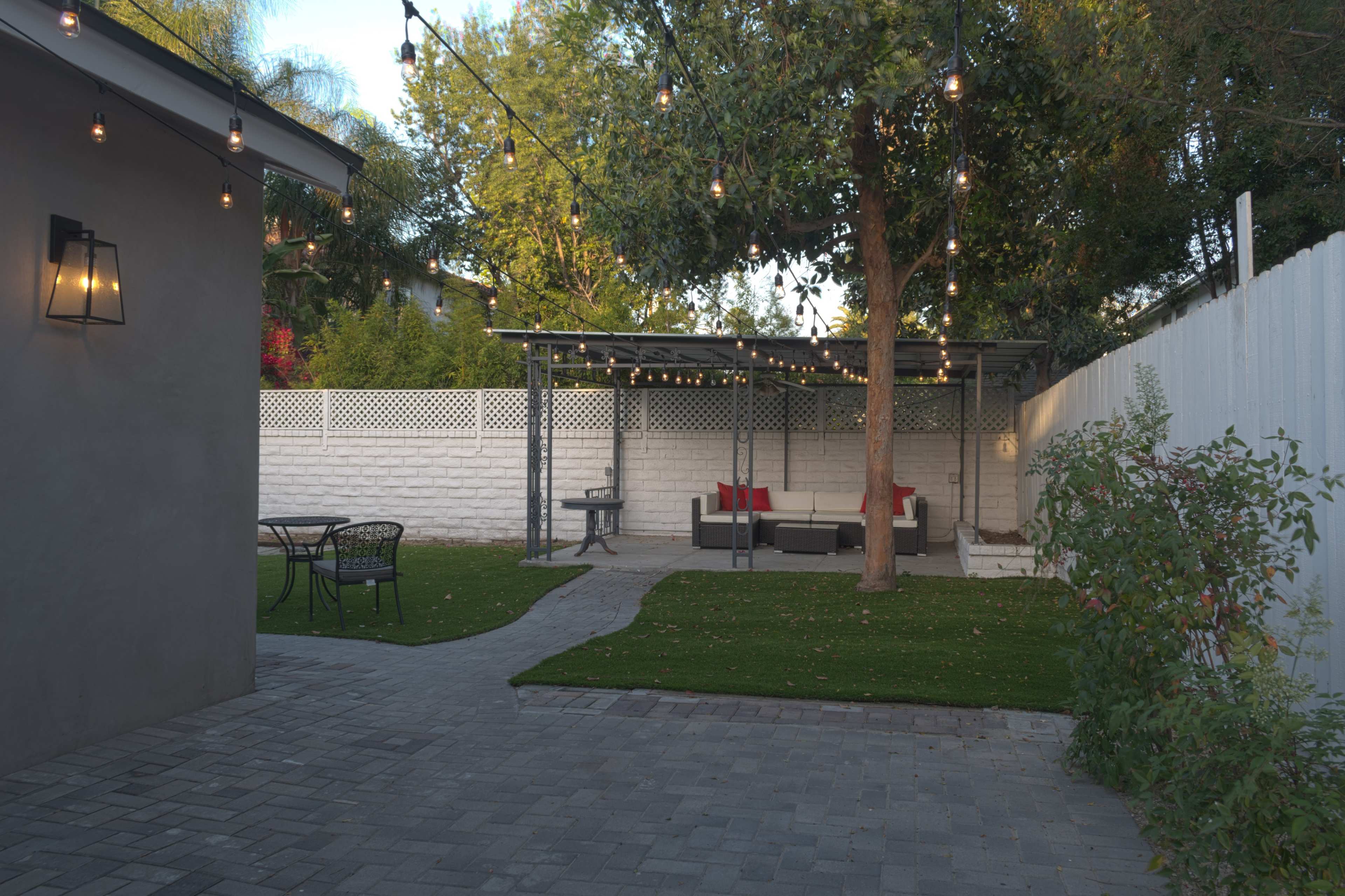 The scene features a well-organized backyard with a seating area under string lights, divided by a stone pathway and surrounded by greenery.