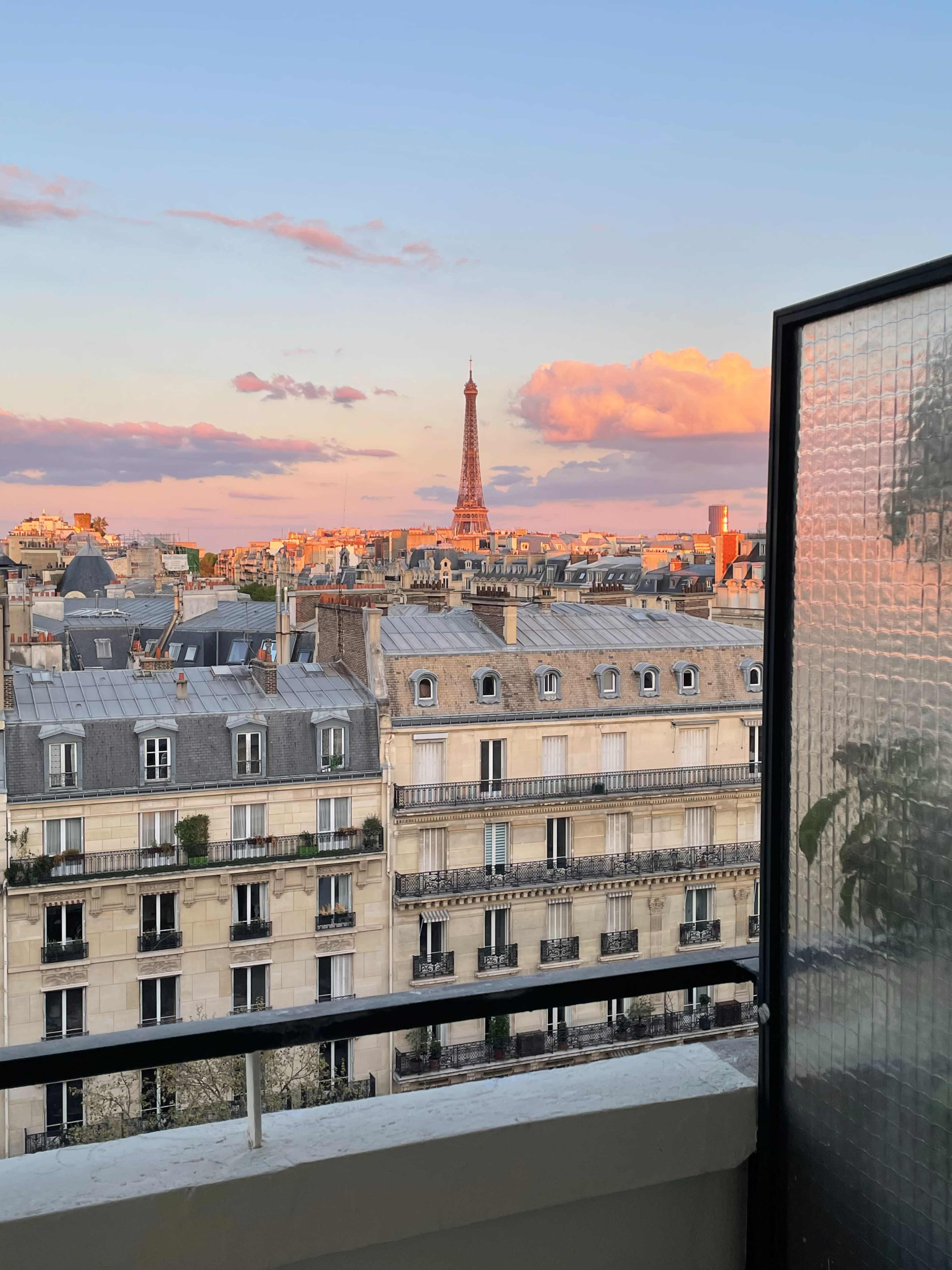 The image shows a view from a balcony overlooking Paris, with the Eiffel Tower visible in the distance against a colorful sunset sky.